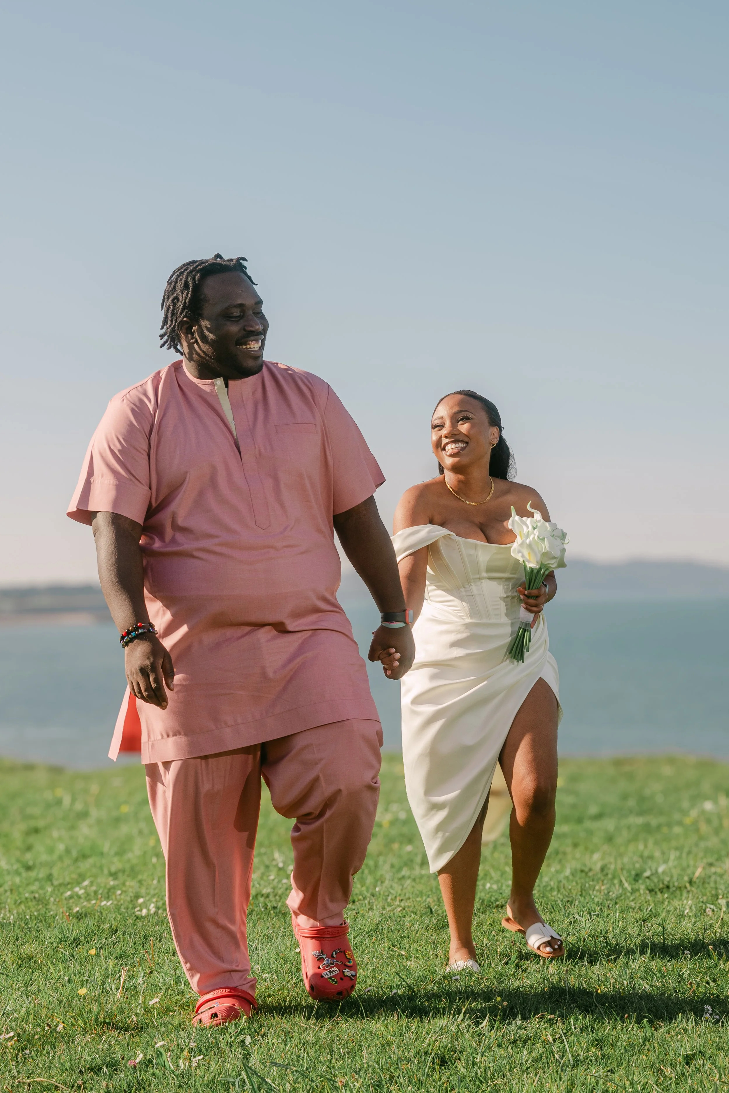 A joyful couple holding hands and walking on grass near a body of water during a sunny day. The man is wearing a pink traditional outfit and sandals, while the woman is in a white dress holding a bouquet.