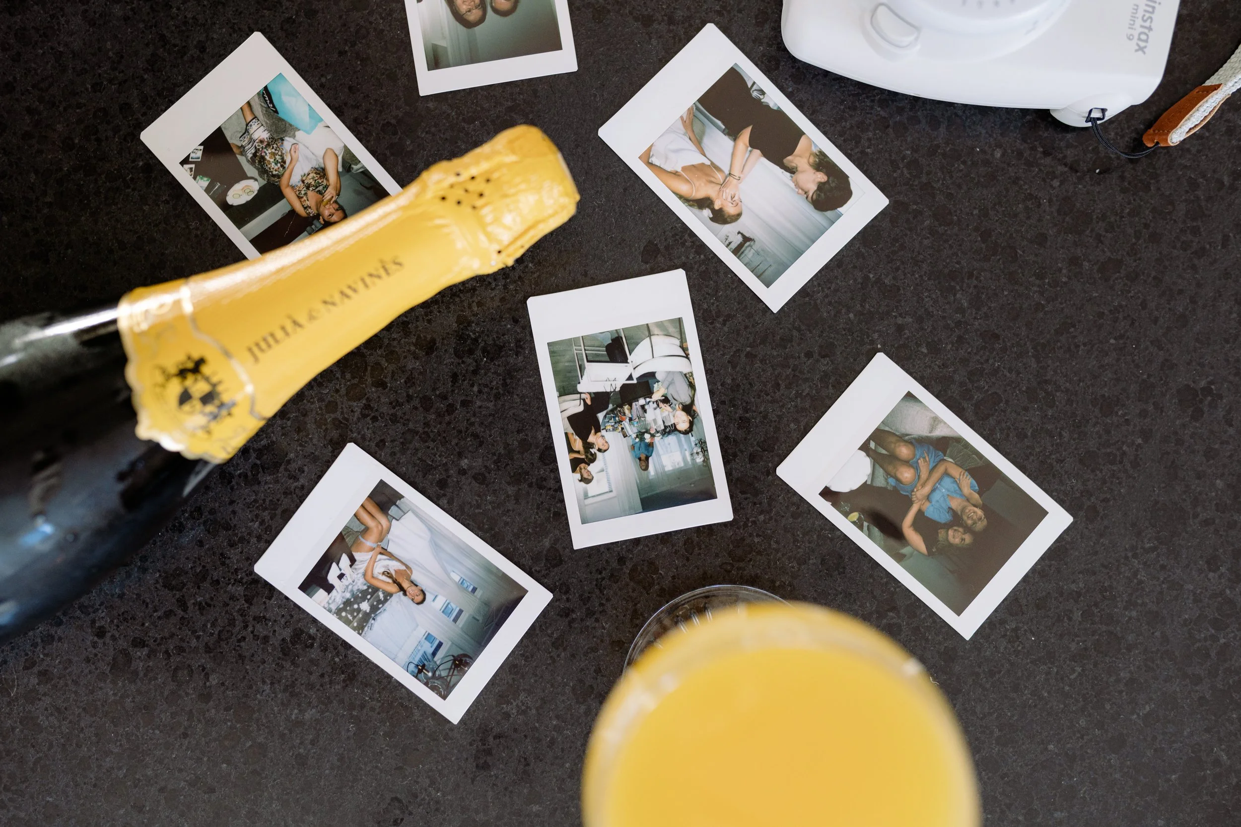 A bottle of Veuve Clicquot champagne lying on a black surface, surrounded by scattered photo booth pictures of people at a social gathering, with a glass of orange juice in the foreground.