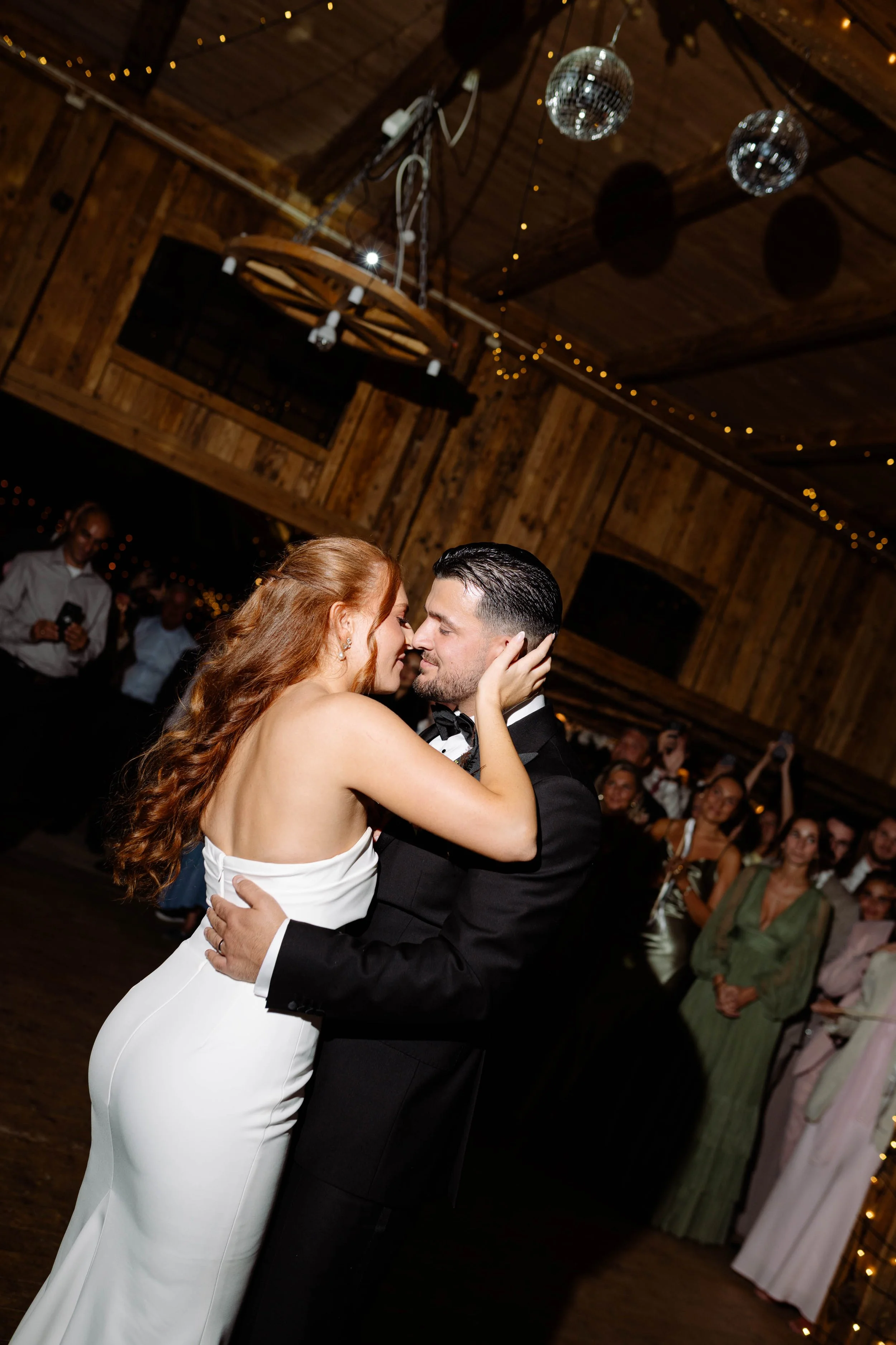 A bride and groom share a dance at their wedding reception in a rustic wooden venue, with guests watching in the background, and disco balls and string lights hanging from the ceiling.