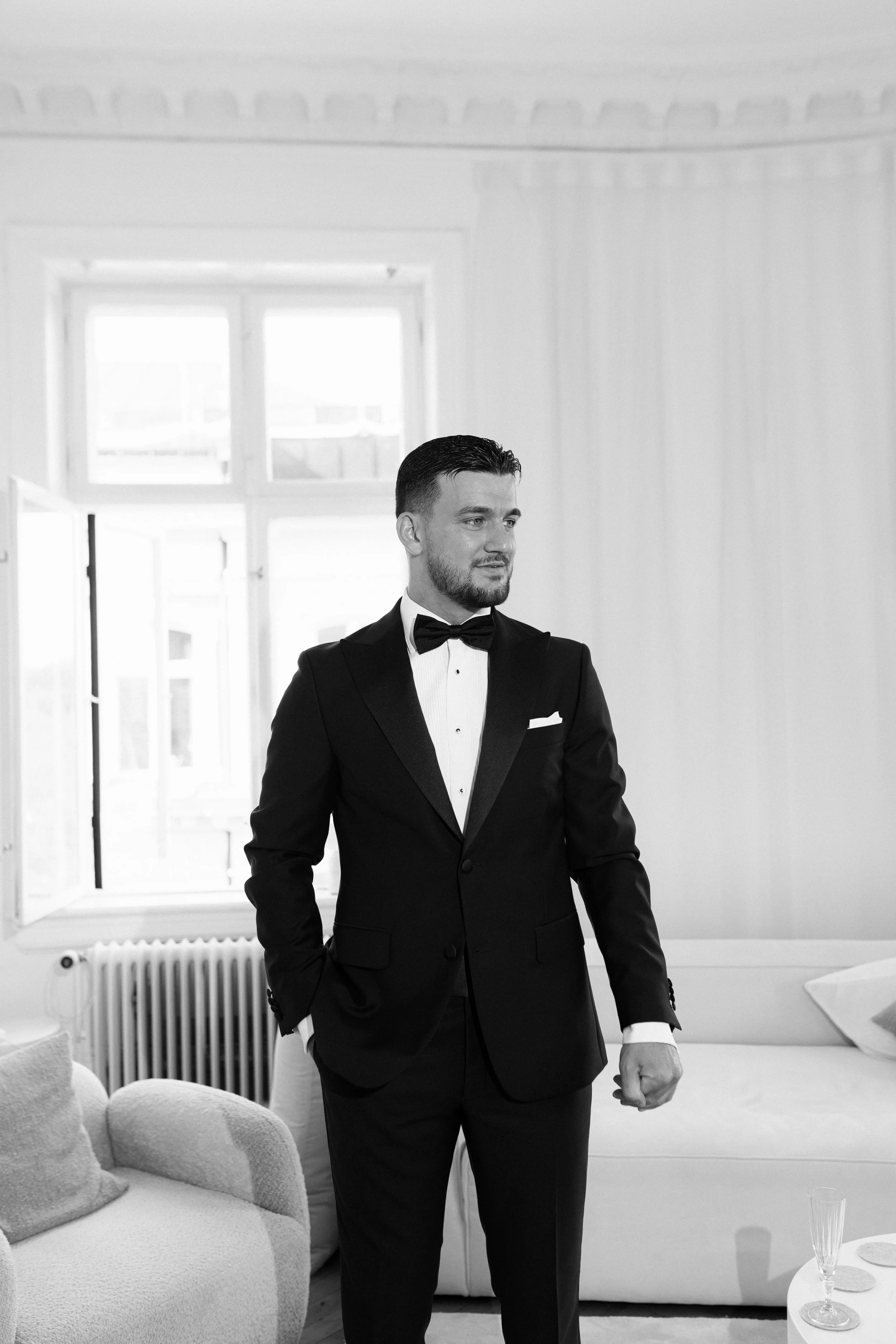 A man in a tuxedo standing in a bright, elegant room with large windows and a sofa.