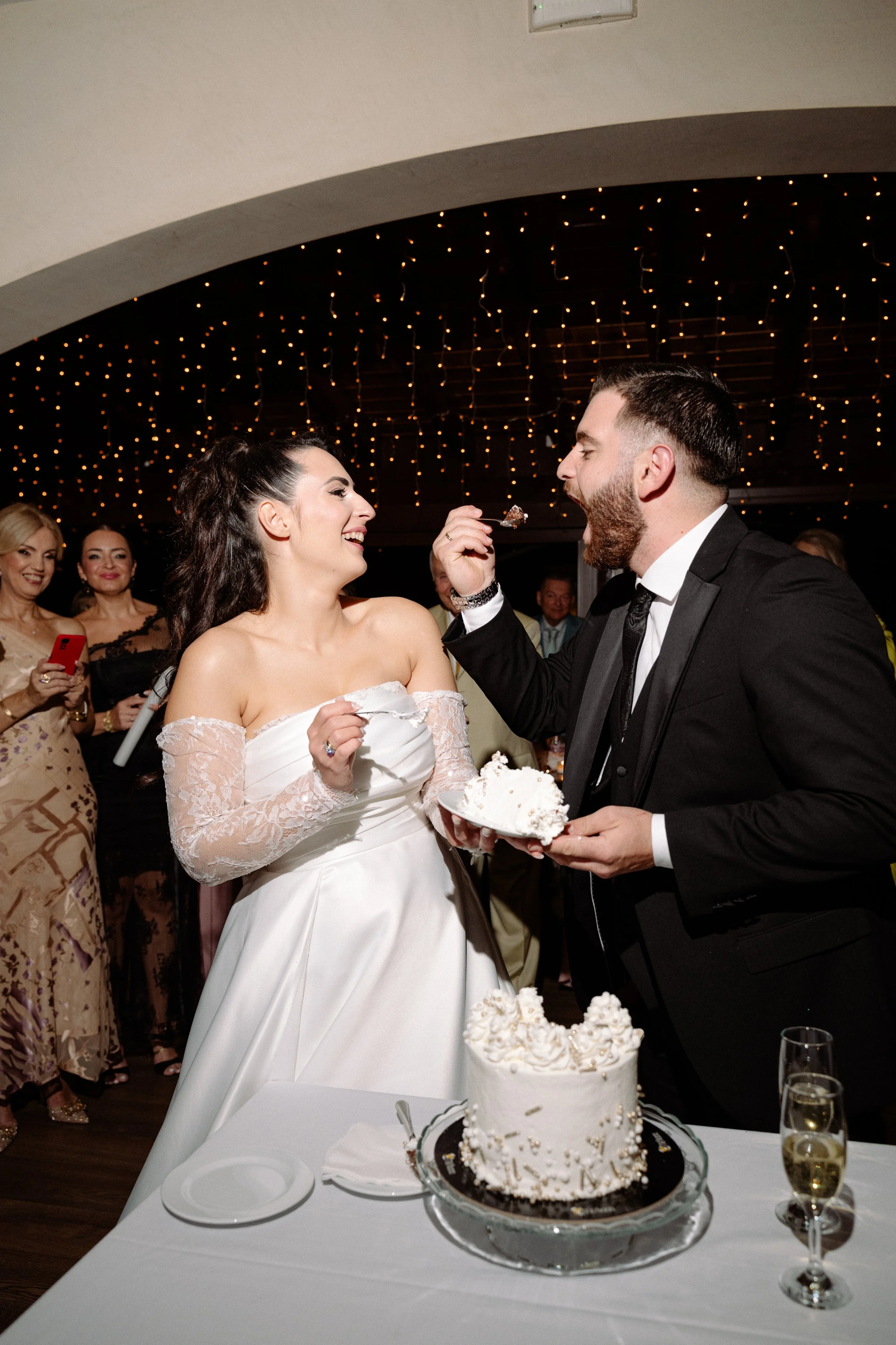 Bride and groom sharing wedding cake at their reception, surrounded by guests, with a decorated ceiling of string lights.