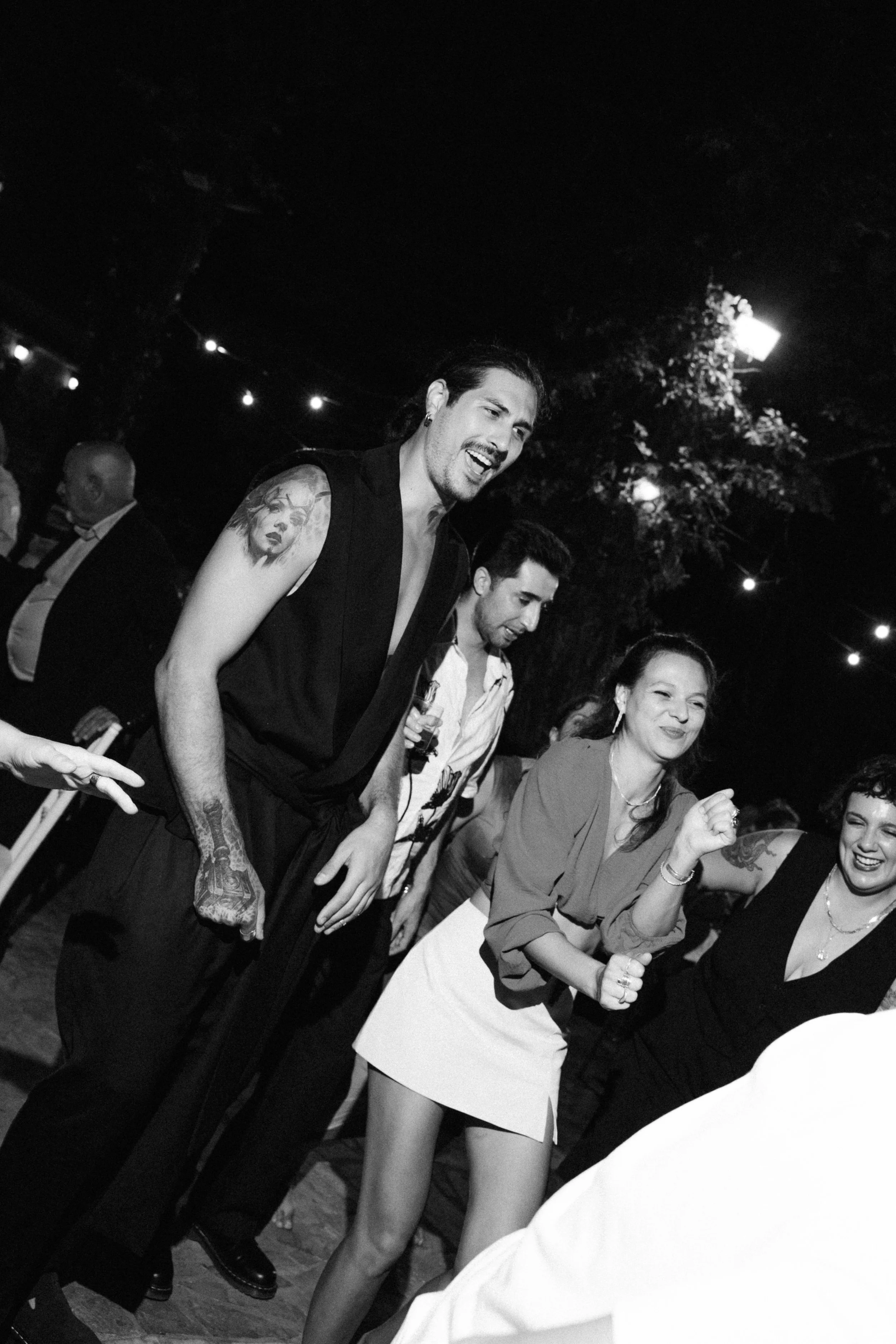 People dancing and socializing at an outdoor night event, with string lights overhead.