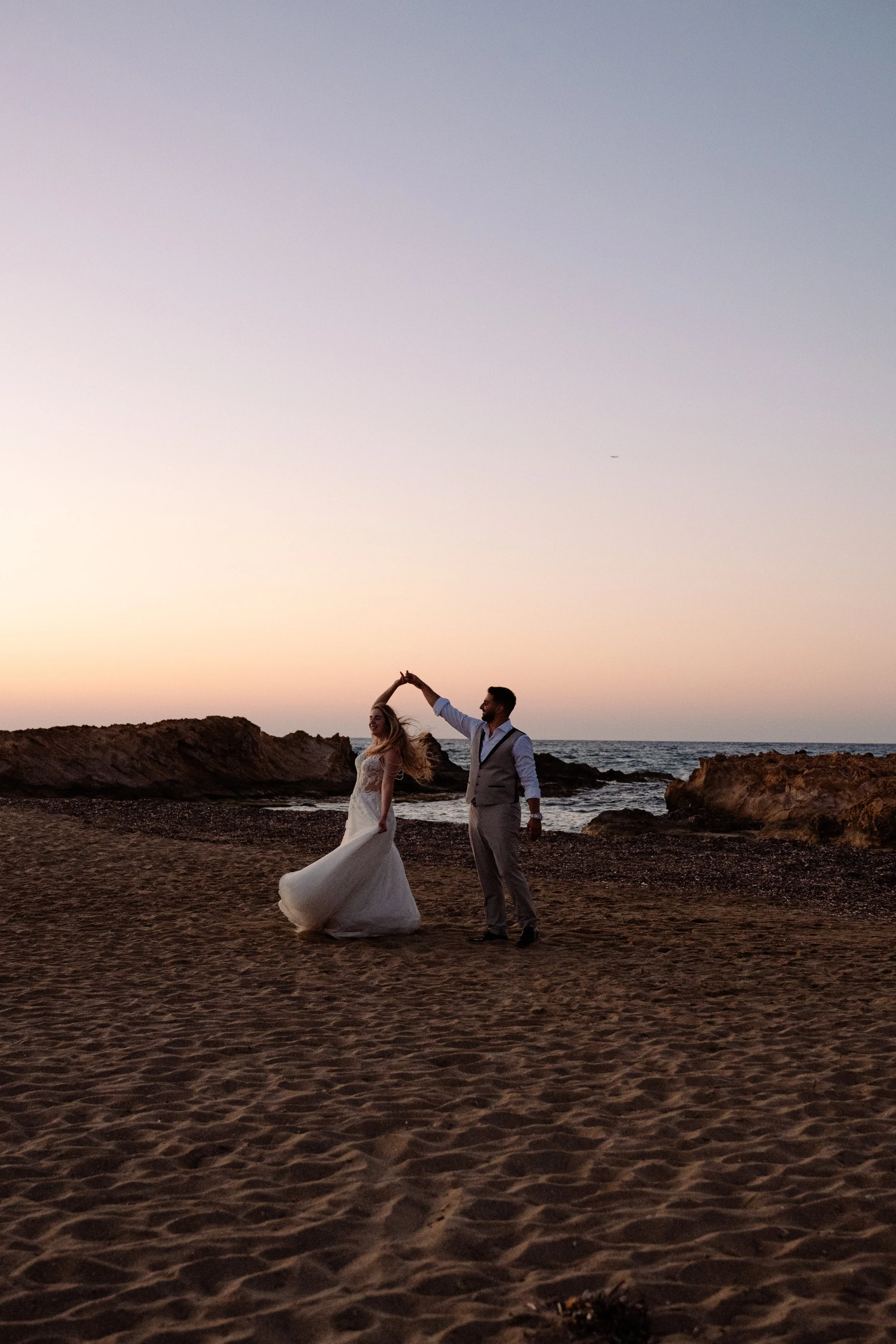 A couple in wedding attire dancing on the beach at sunset, with the groom twirling the bride as she spins in her white wedding dress.
