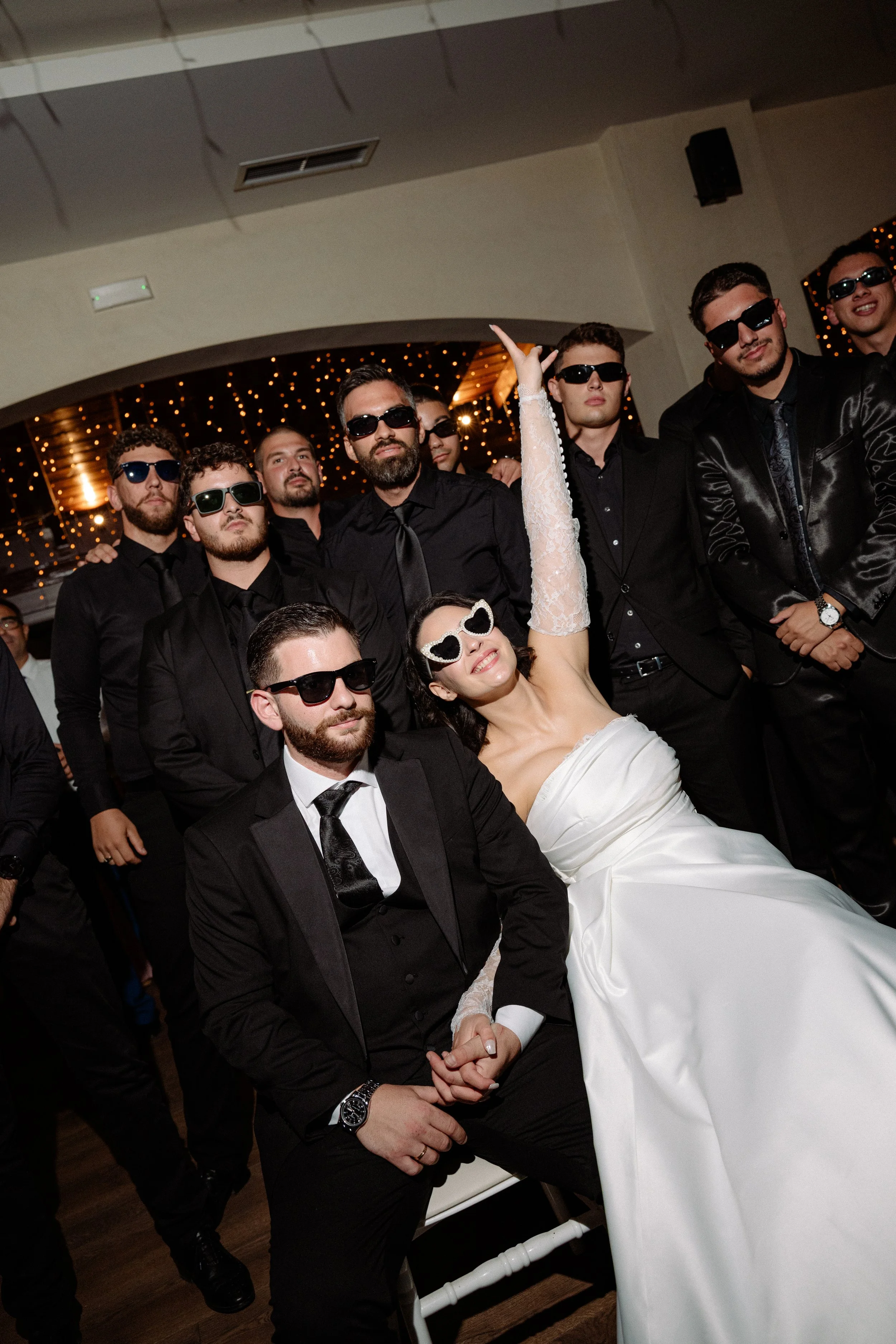 A group of men and a woman in a wedding dress, all wearing sunglasses, at a wedding reception, with string lights in the background.