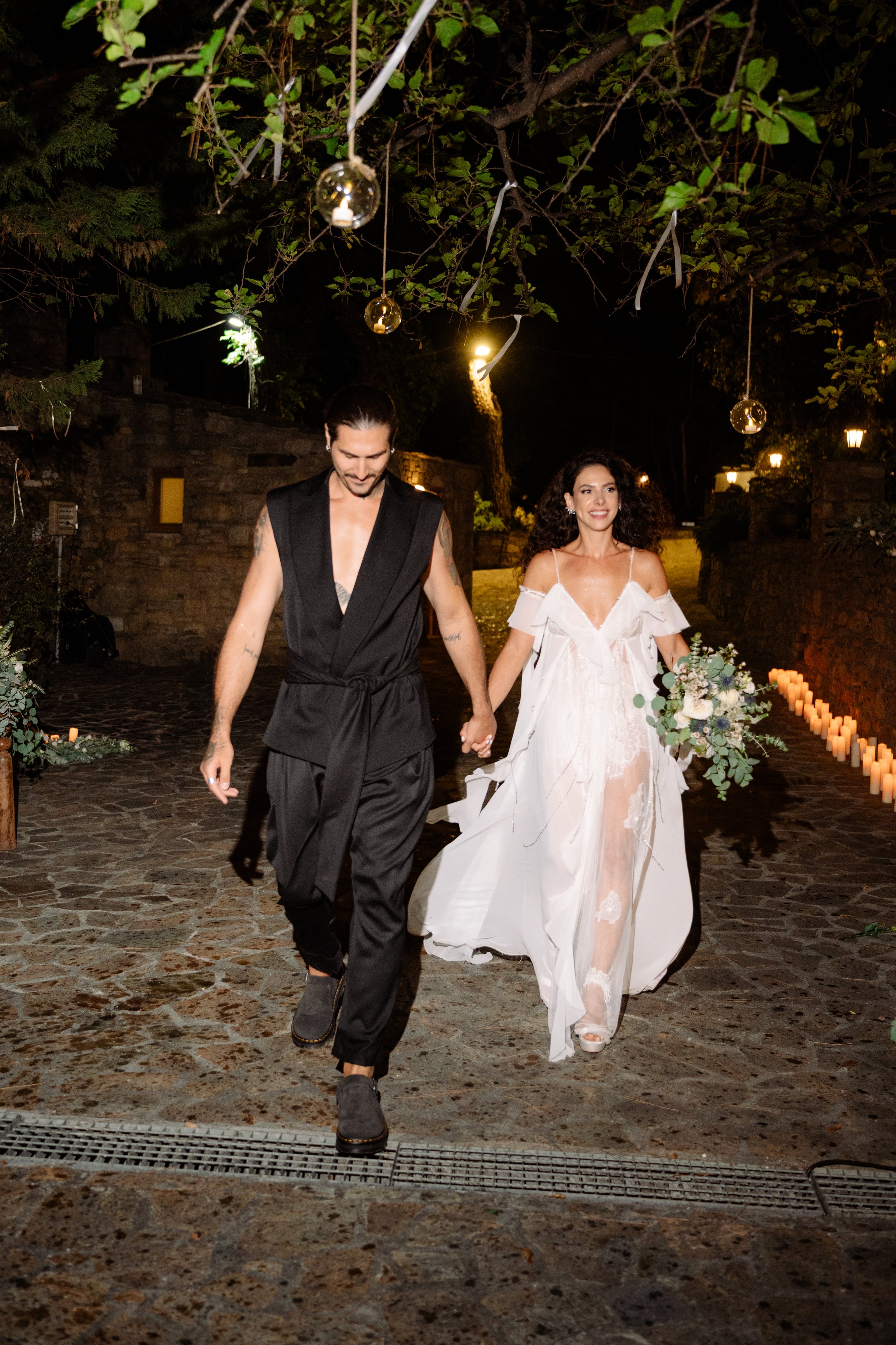A couple holding hands and walking outdoors at night, with the woman wearing a white wedding dress and holding a bouquet, and the man dressed in black casual attire surrounded by decorative hanging lights and candles along the pathway.