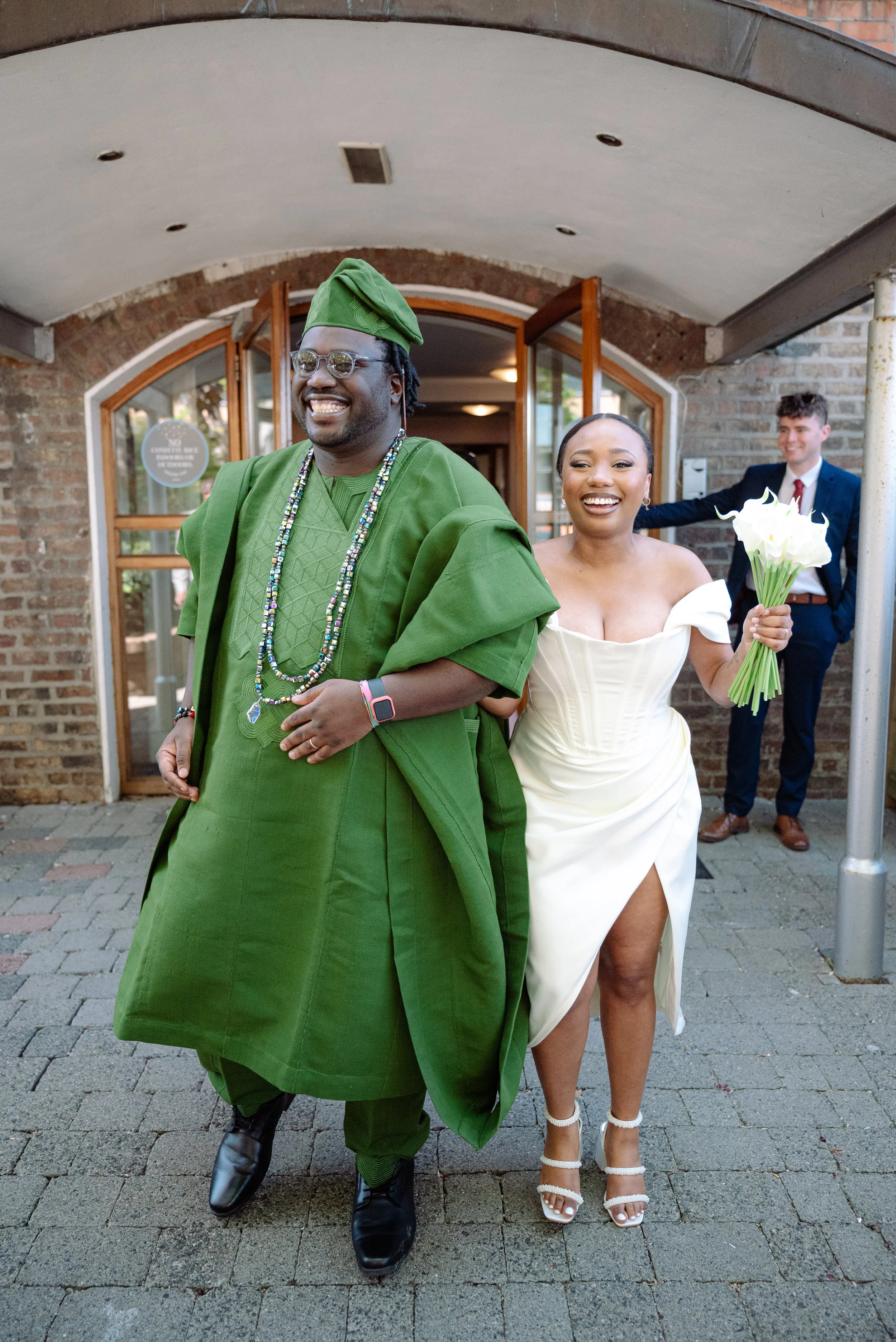 A bride in a white wedding dress holding a bouquet of flowers, smiling, walking arm-in-arm with a man dressed in green traditional attire, outdoors in front of a brick building, with a man in a suit in the background