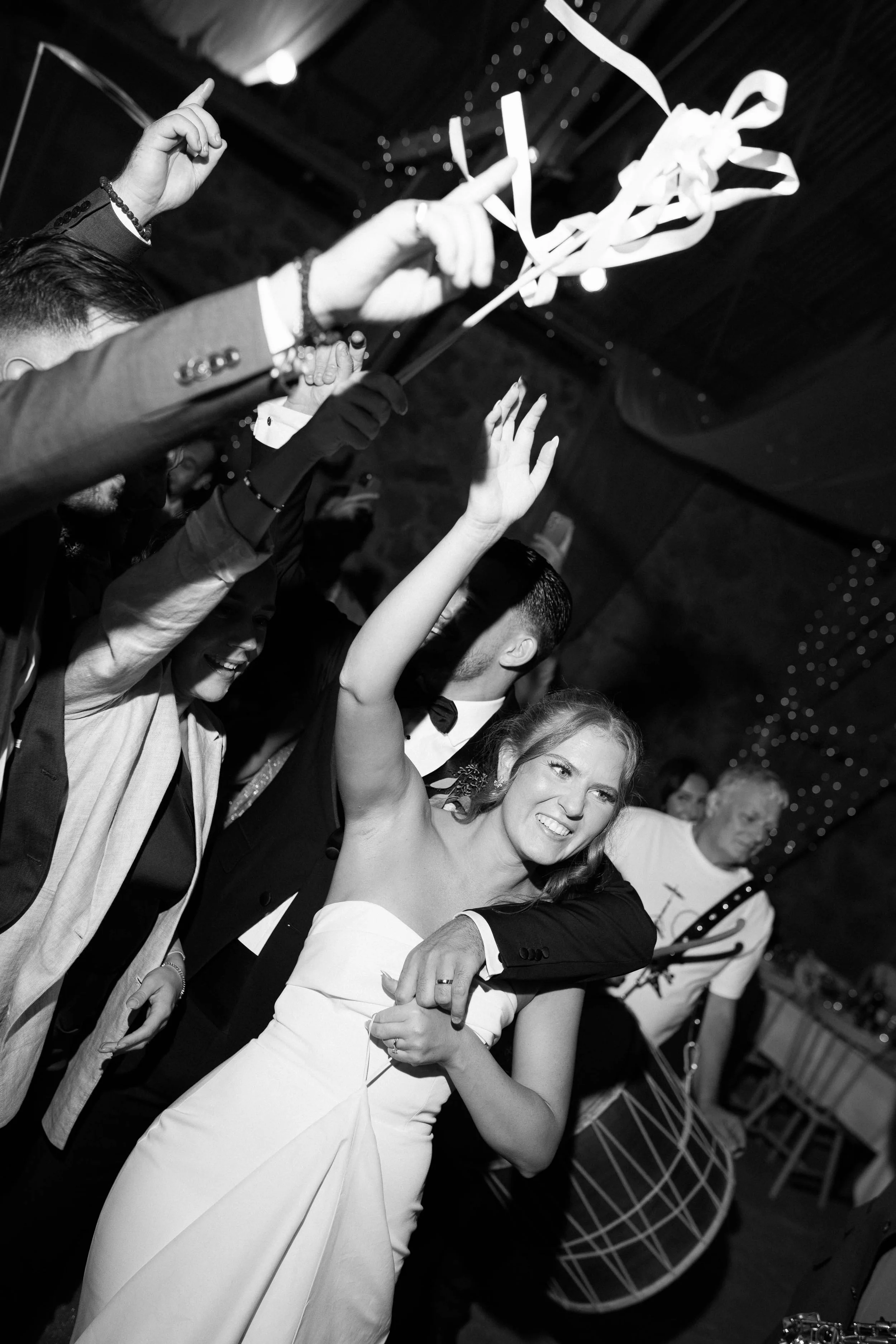 A group of people, including a woman in a white dress, dancing and celebrating at a party or wedding, with someone holding ribbons and streamers overhead.