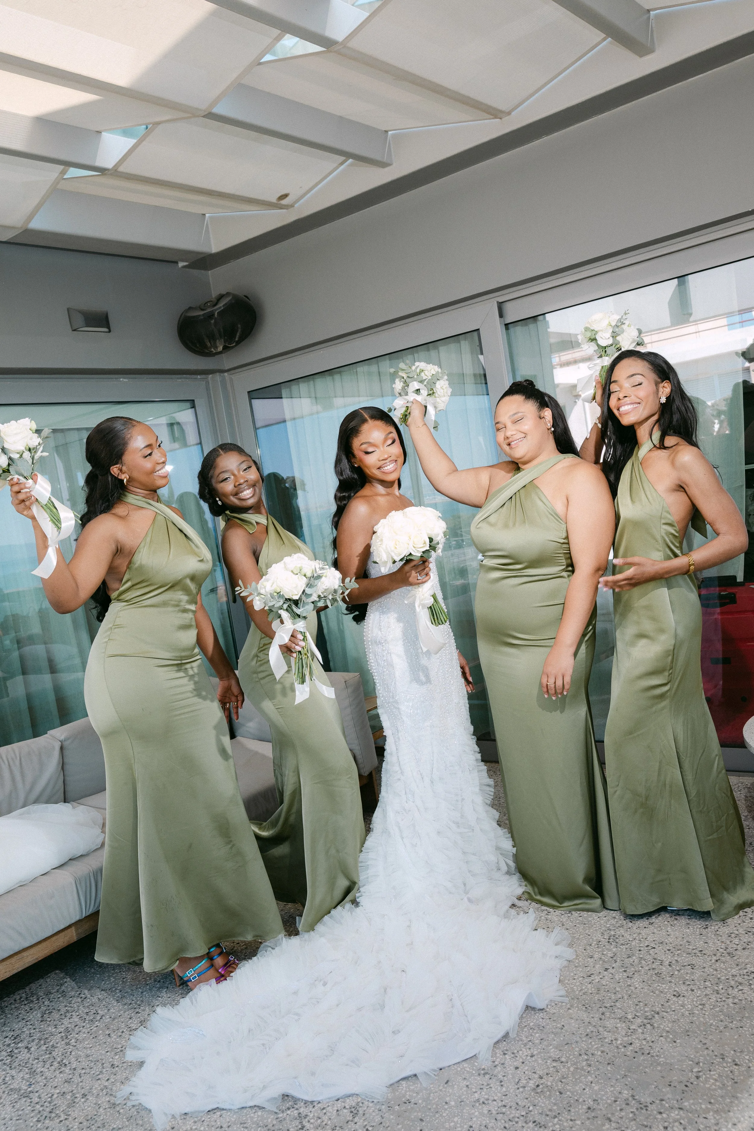 A bride and her five bridesmaids celebrating indoors, with the bridesmaids wearing matching satin olive green dresses and the bride in a white wedding gown, holding bouquets of white roses, smiling and laughing.