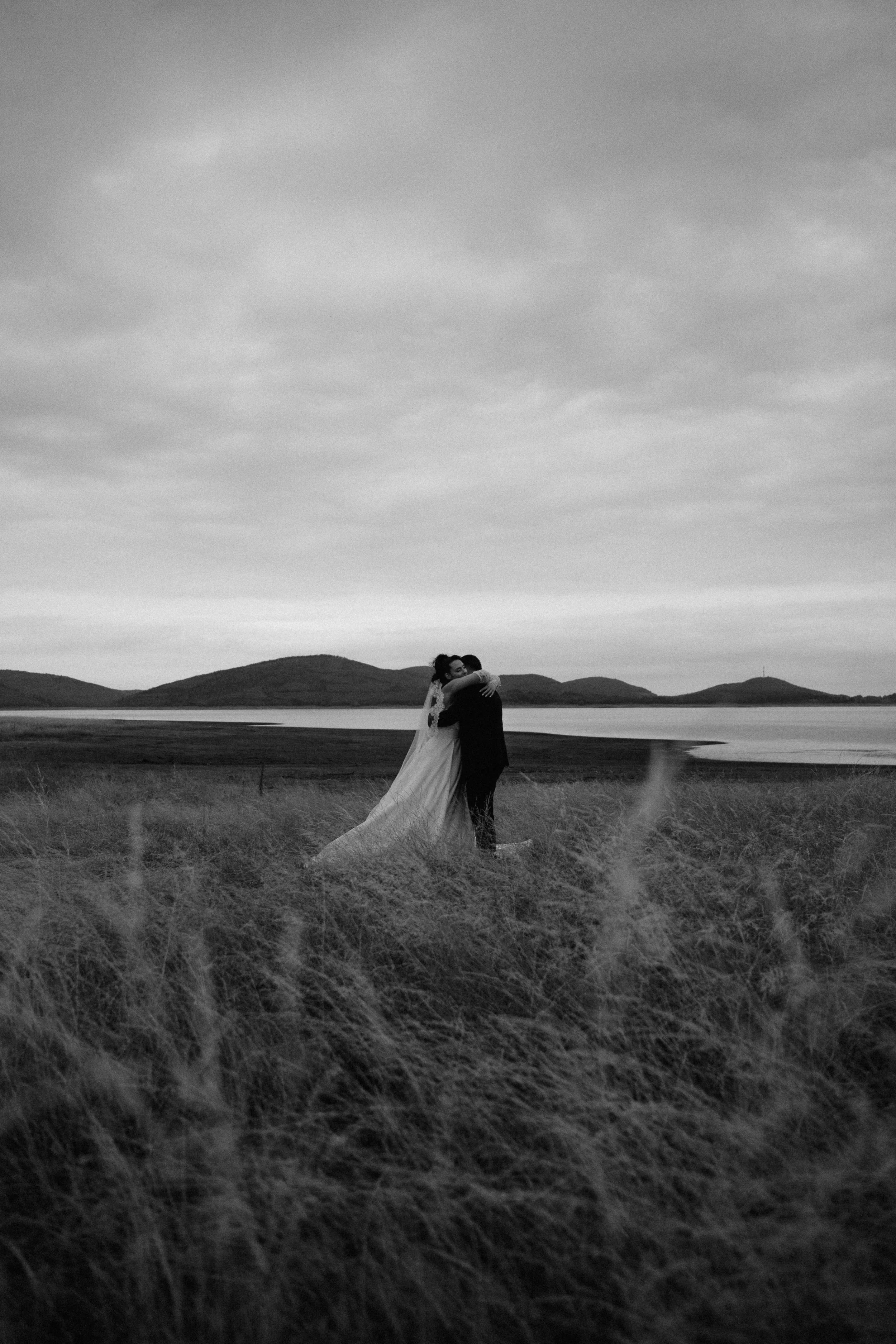 Black and white photo of a bride and groom hugging on a grassy field with a lake and hills in the background.