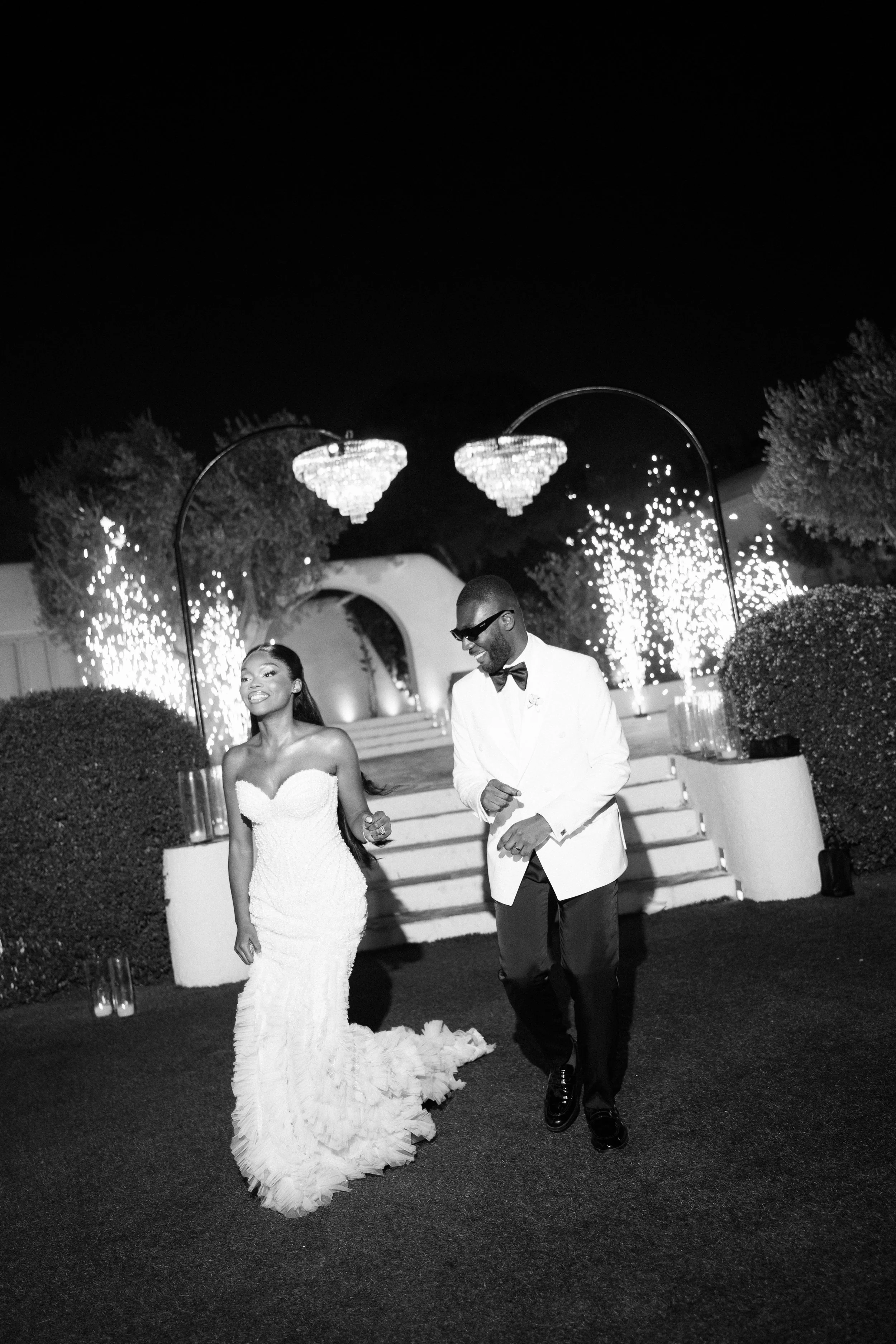 Black and white photo of a bride and groom dancing outdoors at night, with fireworks and chandeliers in the background.