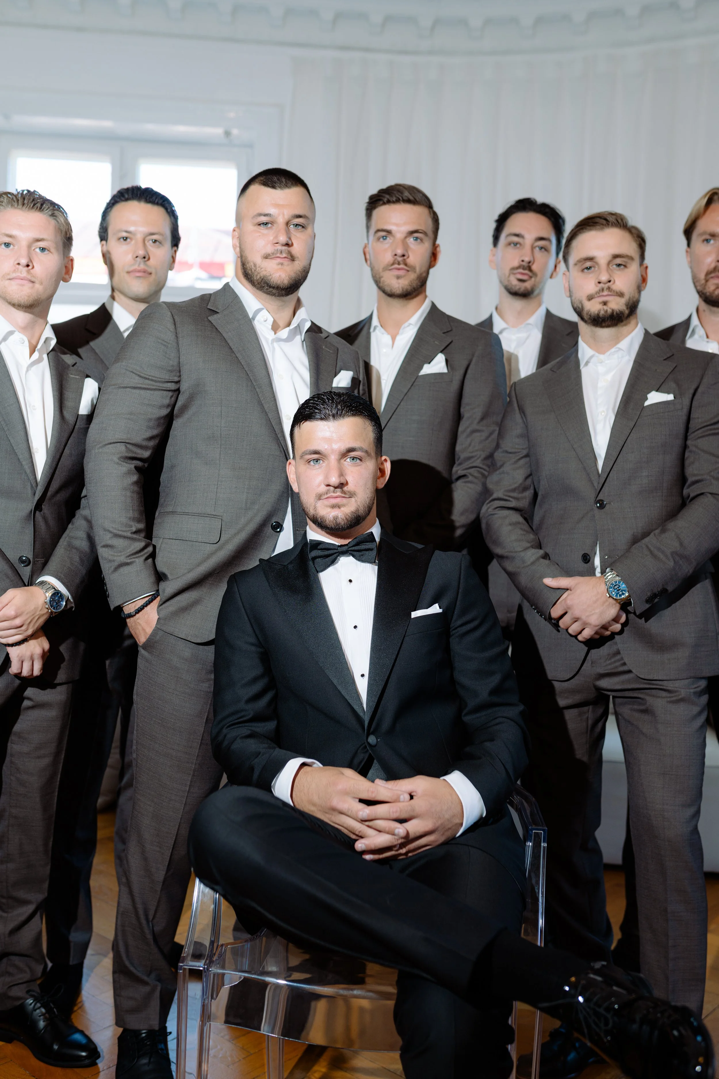A group of men dressed in suits, with one man in the front seated in a black tuxedo with a bow tie, all posing for a formal group photo indoors.
