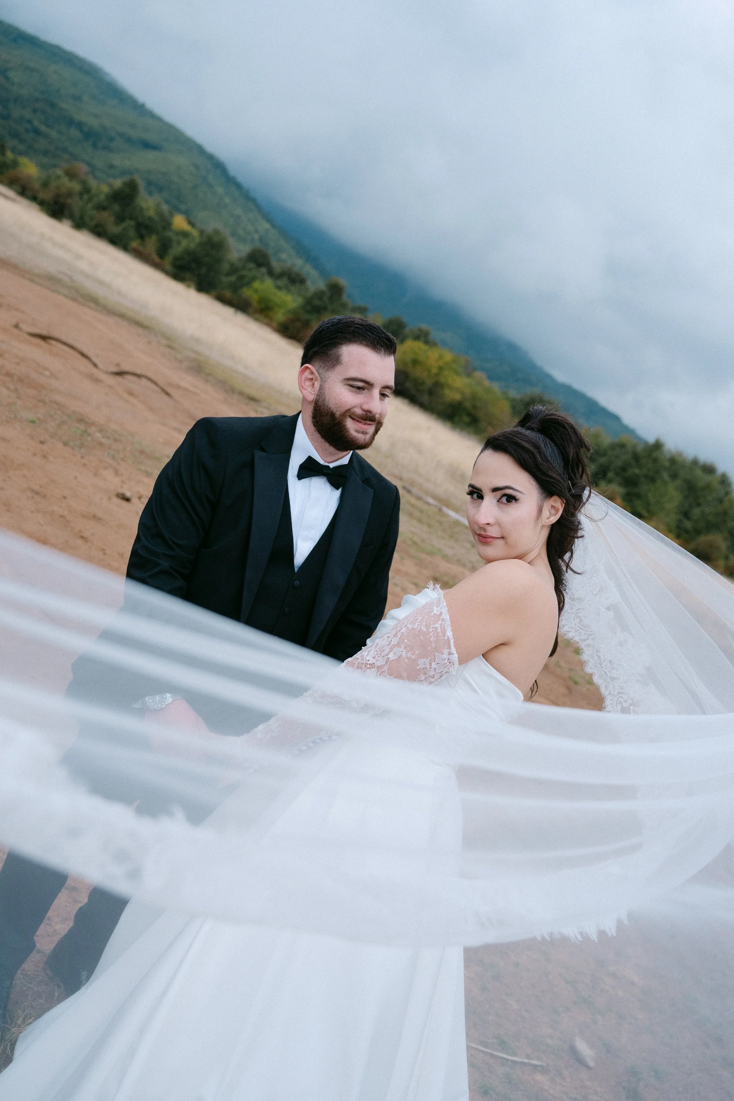 Bride and groom in wedding attire outdoors with mountains in the background, overcast sky, the bride's veil flowing in the breeze.