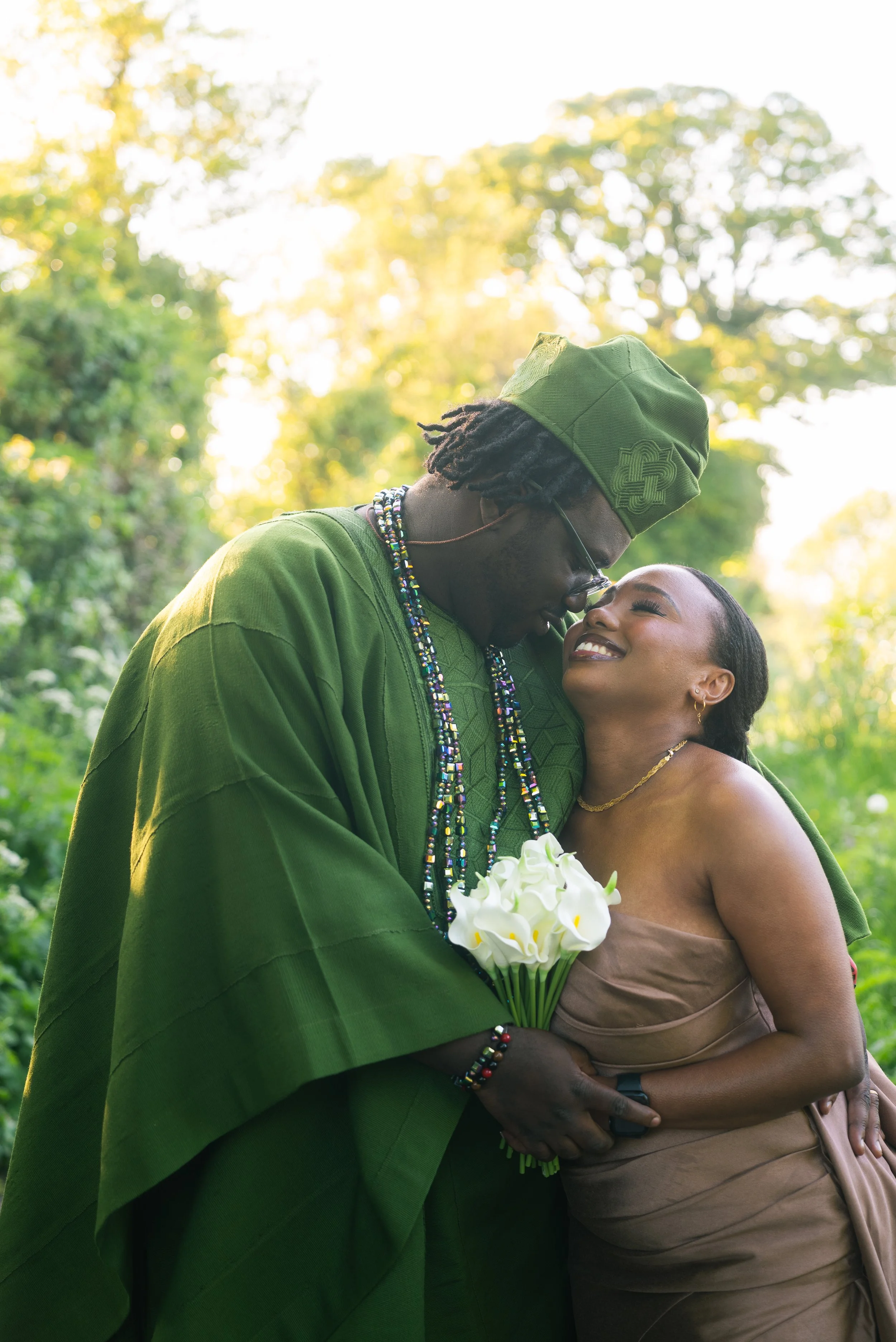 A couple is embracing outdoors with sunlight filtering through trees in the background. The man is wearing a green traditional outfit and headpiece, and the woman is in a strapless beige dress, holding a bouquet of white calla lilies, and smiling at 