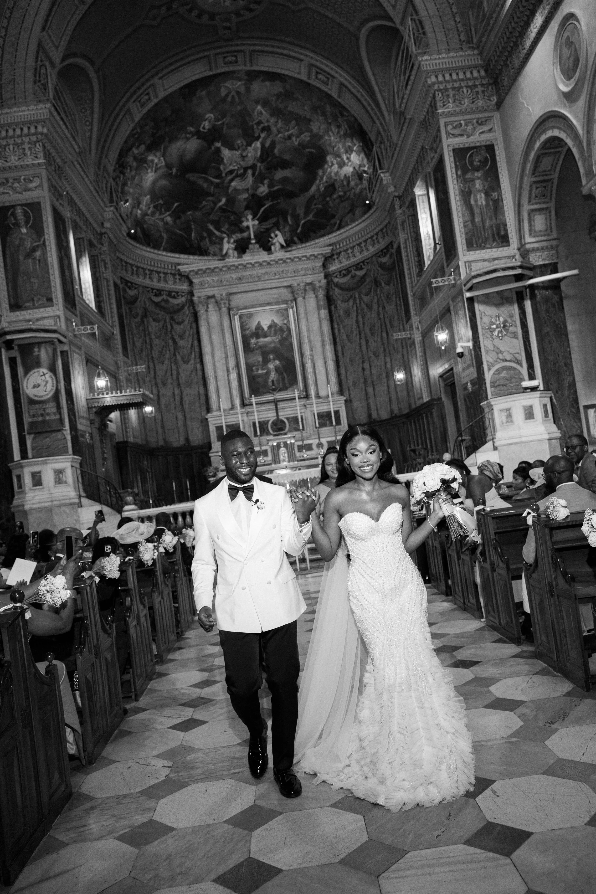 Black and white wedding photo of a bride and groom walking down the aisle inside a church, with guests seated on either side. The bride is holding a bouquet and wearing a strapless wedding gown, while the groom is wearing a suit with a bowtie. The ch