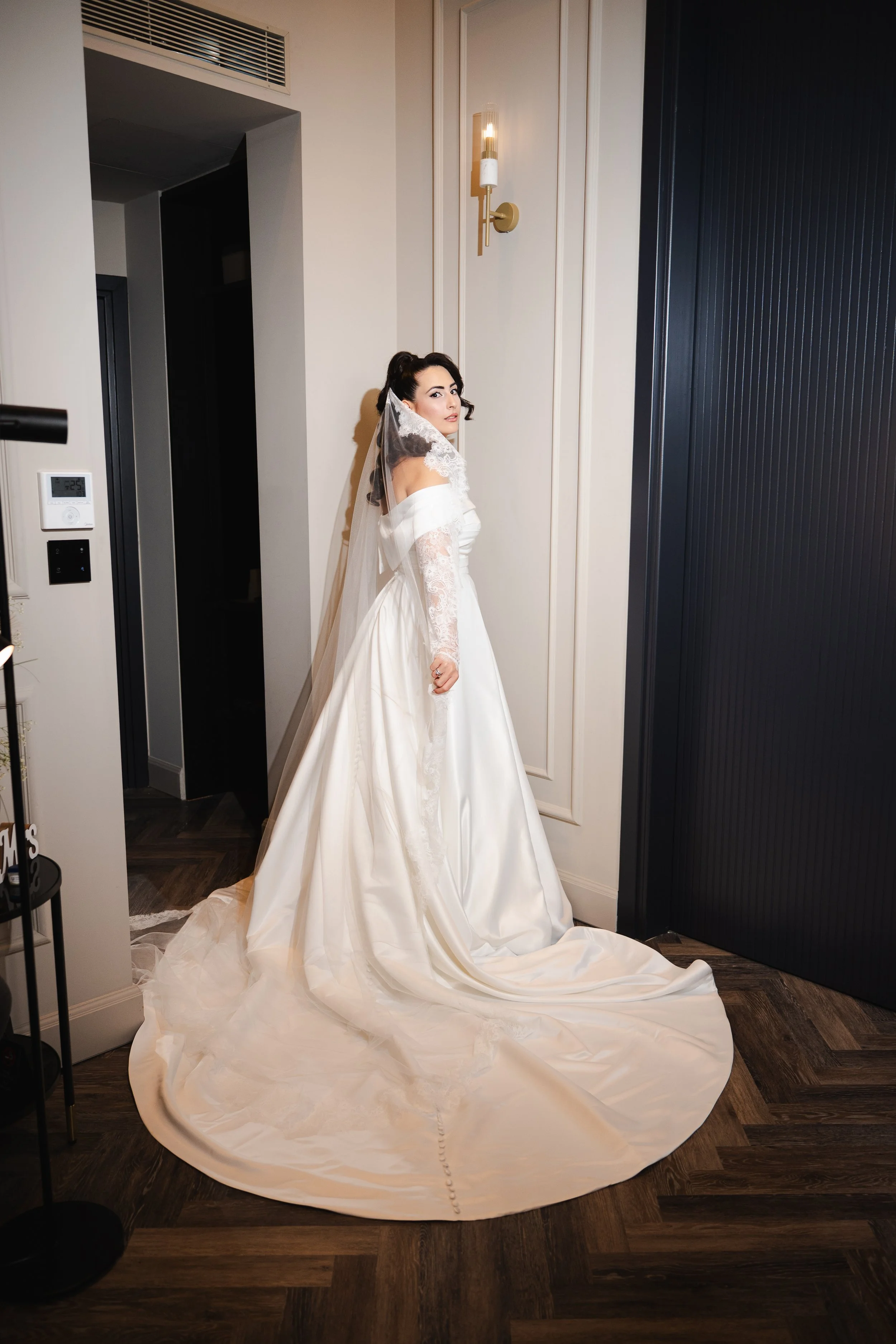 A bride in a white wedding dress and veil stands in a modern, stylish room with dark wood flooring, white walls, and a black door. She looks over her shoulder.