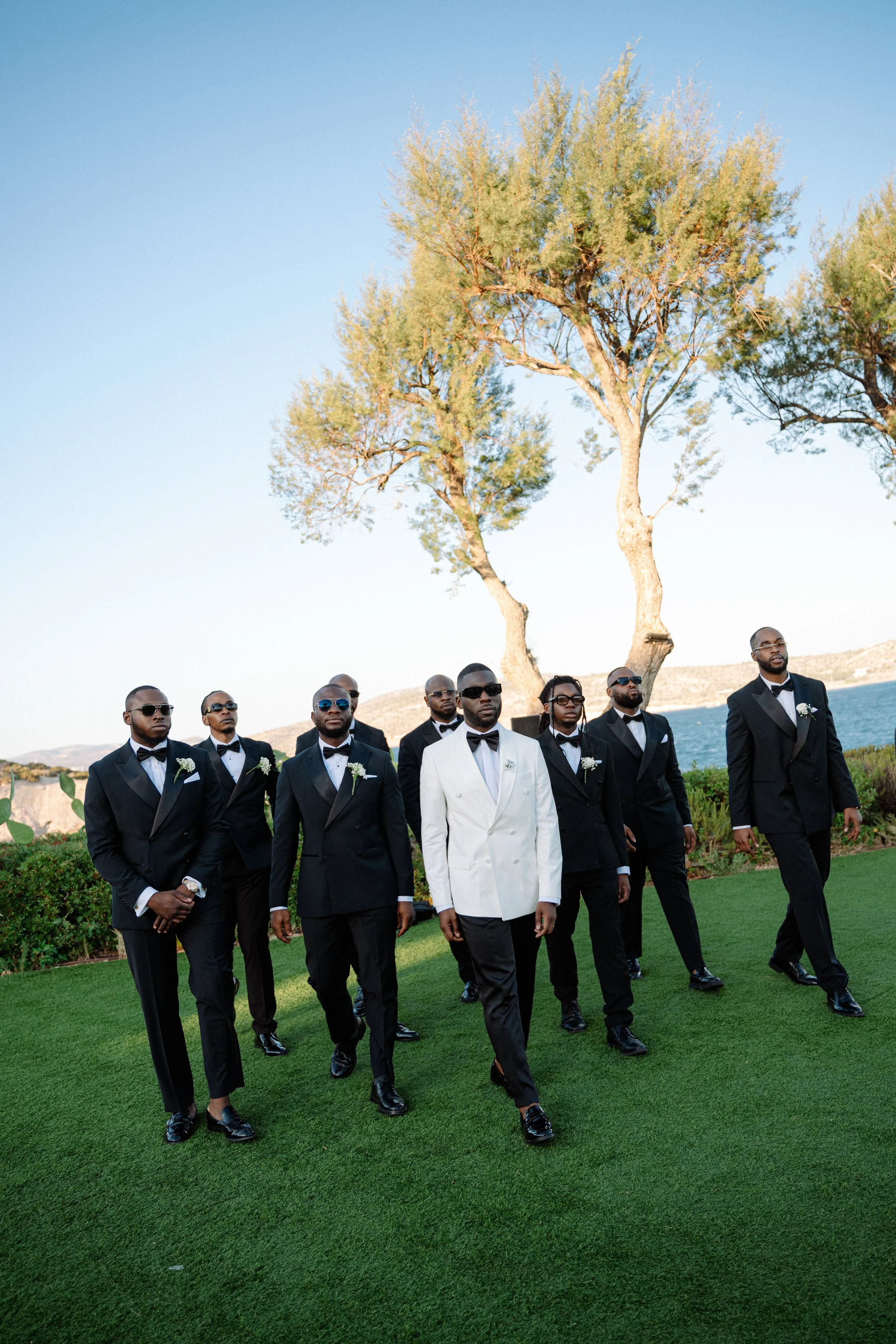 A group of nine men dressed in tuxedos walking on a grassy area near the coast. One man is in a white tuxedo jacket, and the others are in black tuxedos. They are wearing sunglasses and have flowers pinned to their jackets. There are trees and the oc