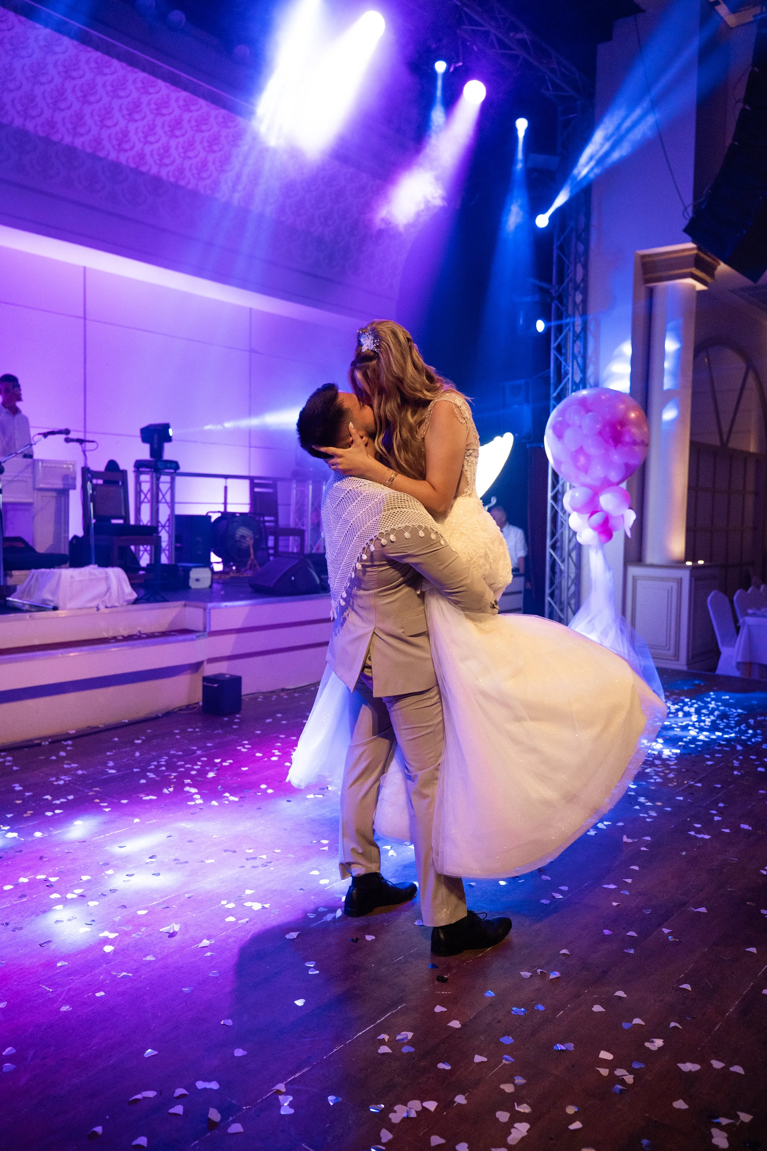 A bride and groom dancing at a wedding reception, with purple and blue lighting, confetti on the floor, and a band setup in the background.