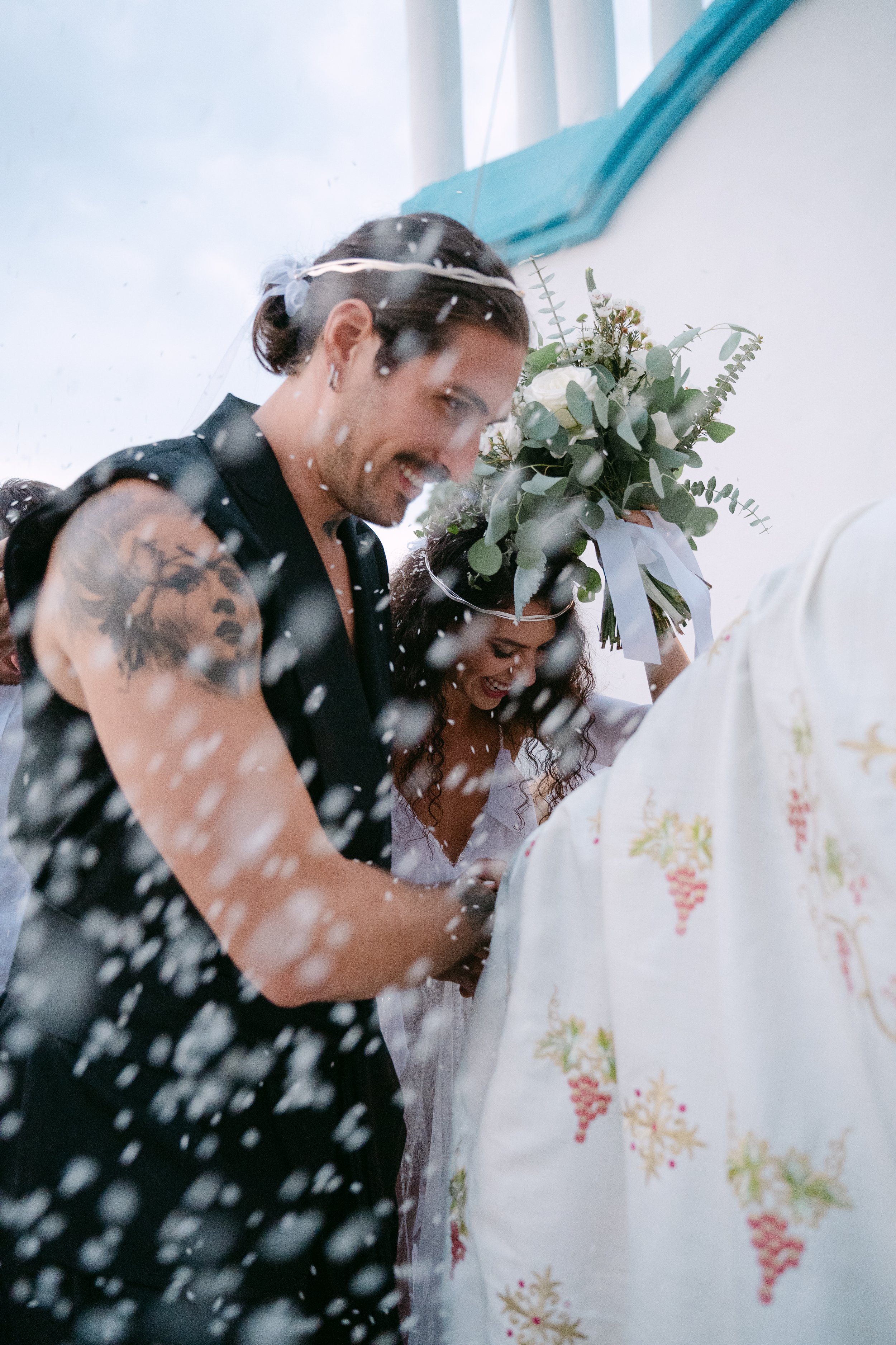 A couple celebrating a wedding ceremony under a spray of water, with the man holding a bouquet of white flowers and greenery, smiling happily.