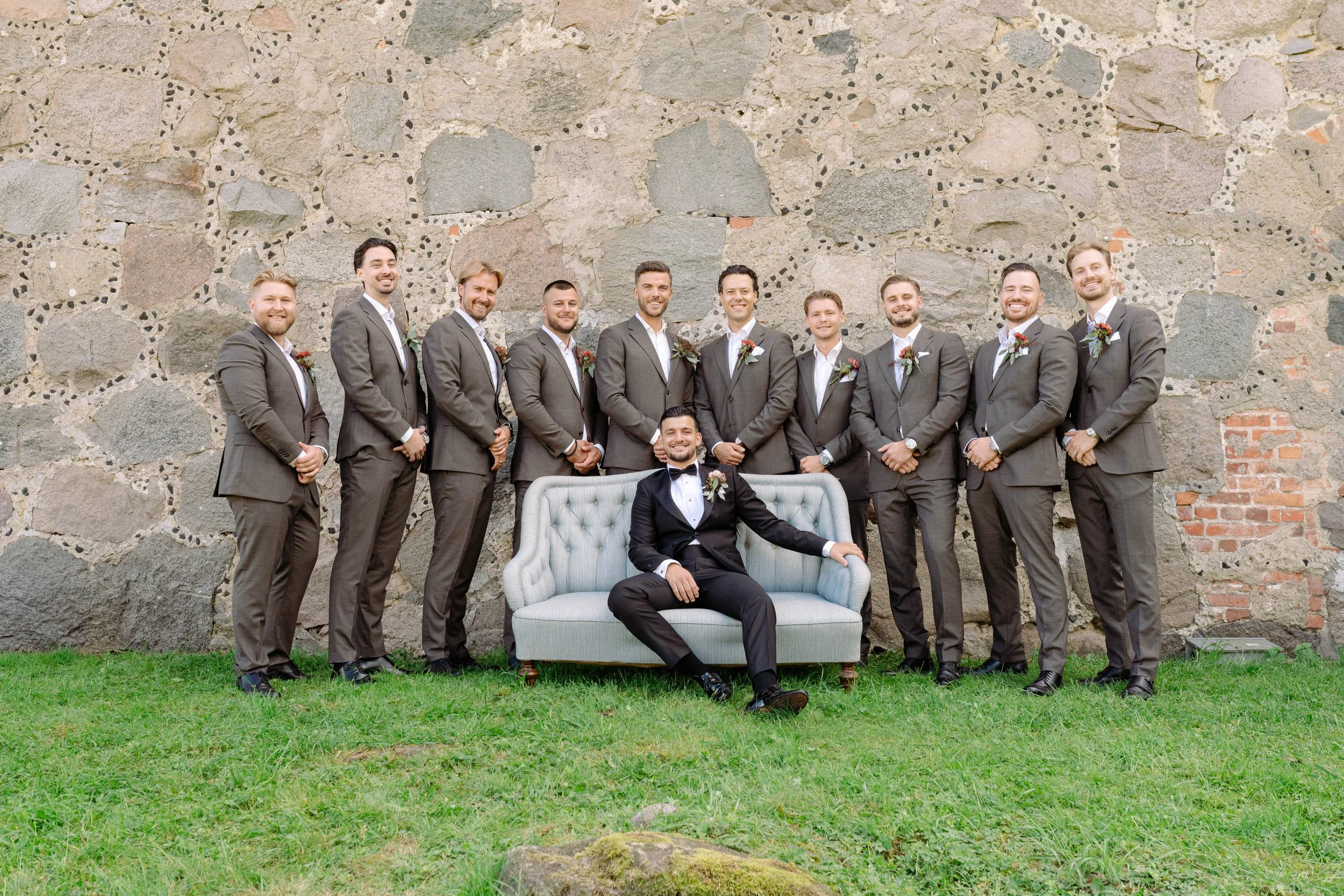 A groom in a black tuxedo sitting on a light-colored vintage sofa in front of a stone wall, surrounded by eleven groomsmen in gray suits with boutonnieres, all smiling.