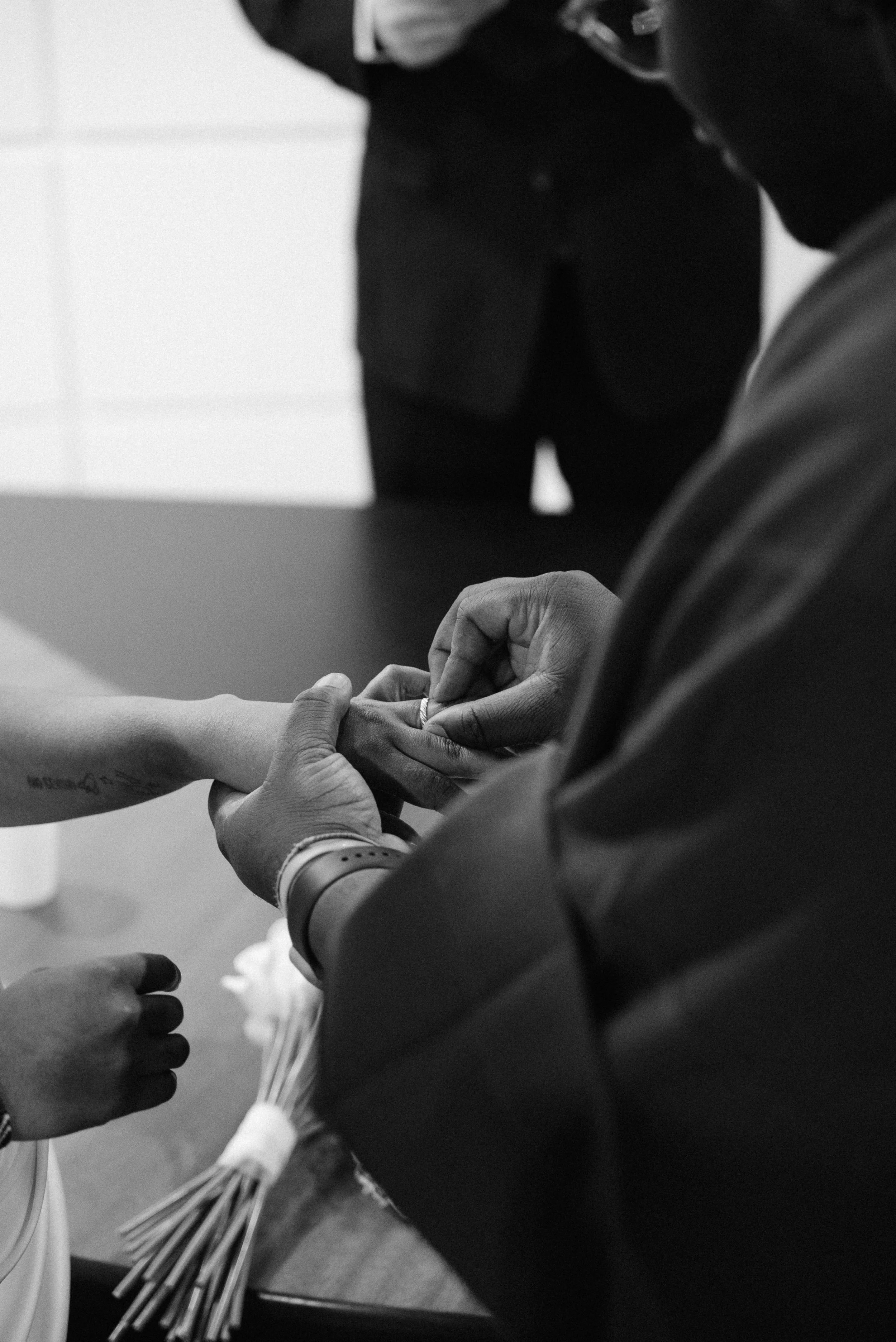 A person is placing a ring on another person's finger during a wedding ceremony with a second person standing in the background.