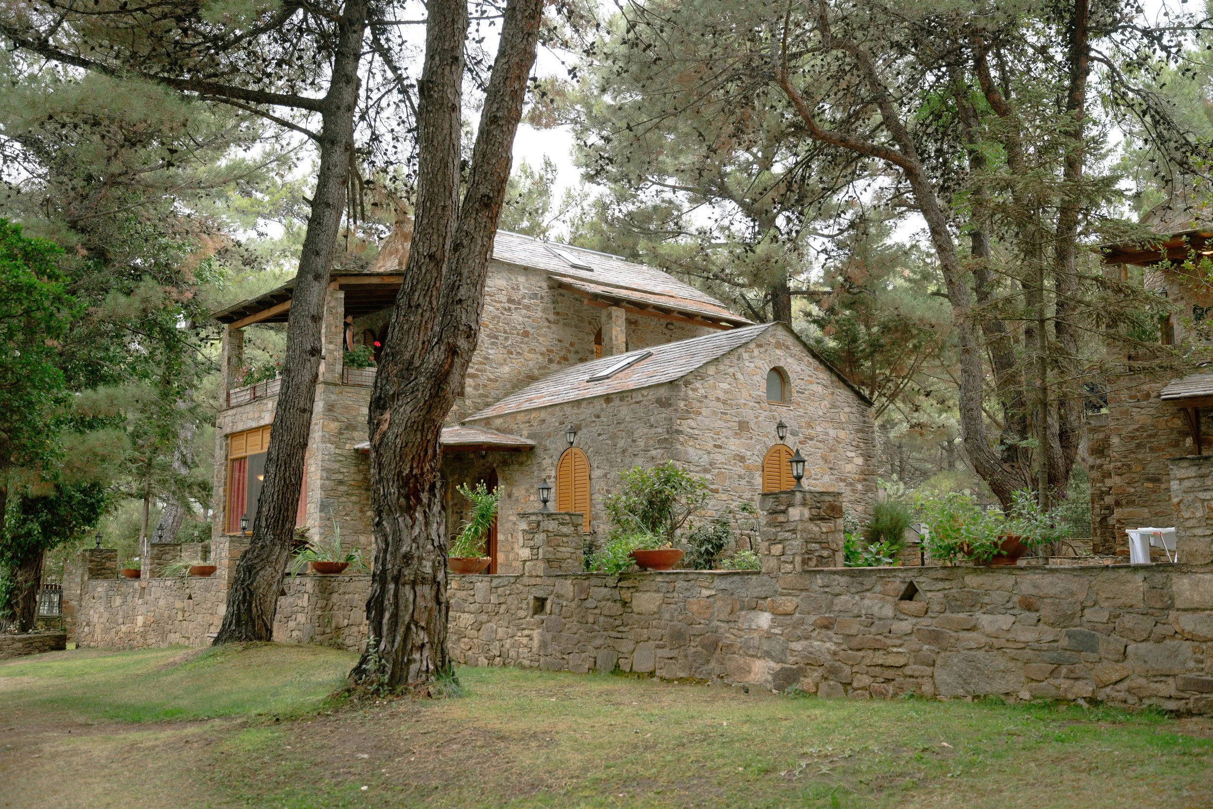 Stone house with multiple levels surrounded by trees and a stone wall, with potted plants and outdoor furniture visible.