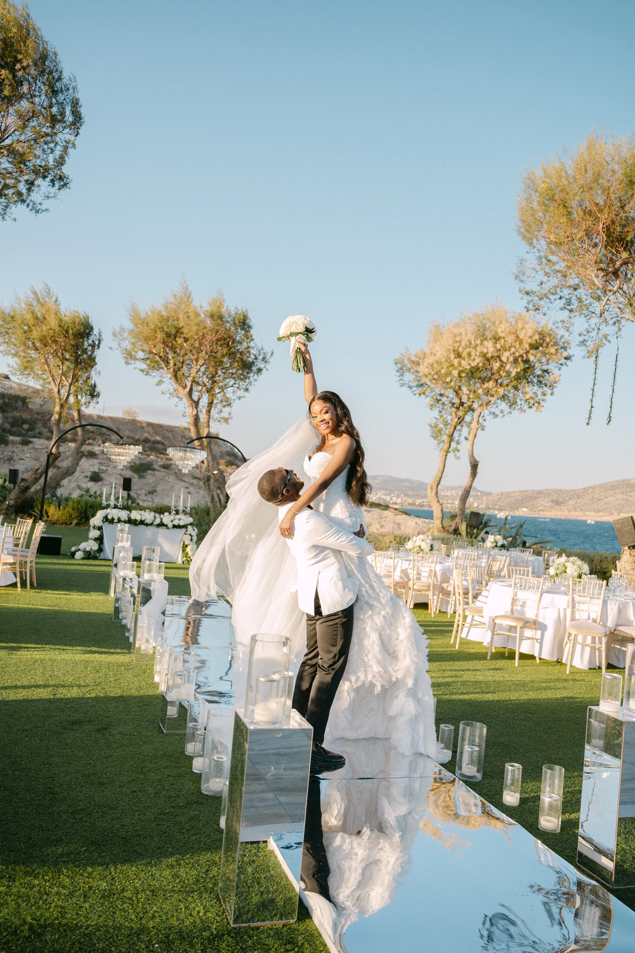 A groom lifts a bride celebrating outdoors at a wedding, with a scenic view of trees, water, and mountains in the background. The bride holds a bouquet and is smiling.