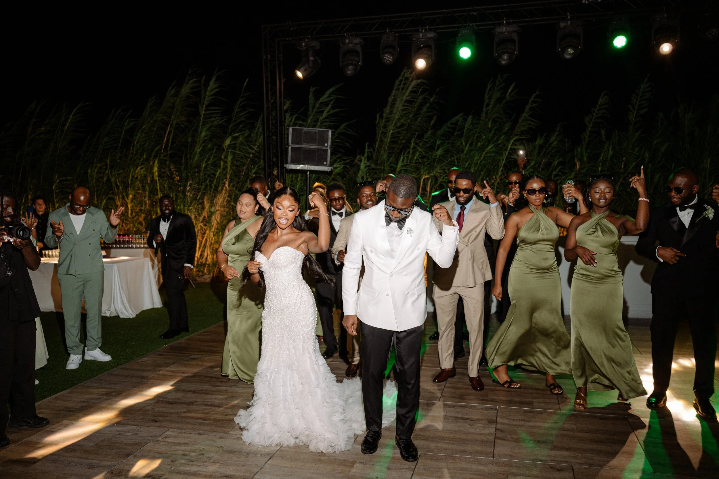 A wedding reception dance floor with a bride in a white gown and groom in a white jacket dancing with guests at night, decorated with plants and illuminated lights.
