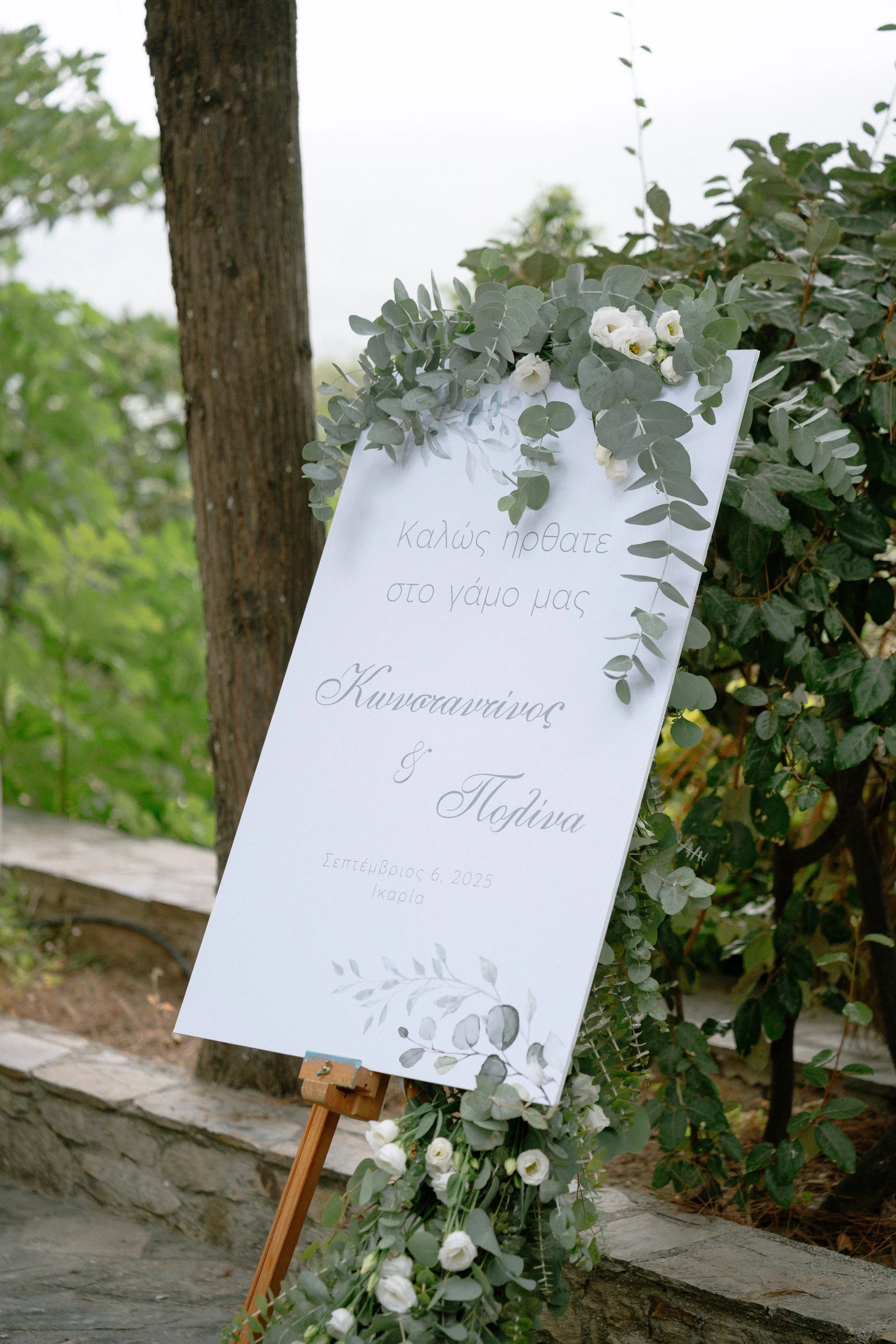 Wedding welcome sign decorated with eucalyptus leaves and white flowers, displaying Greek text and wedding details on a stand outdoors.