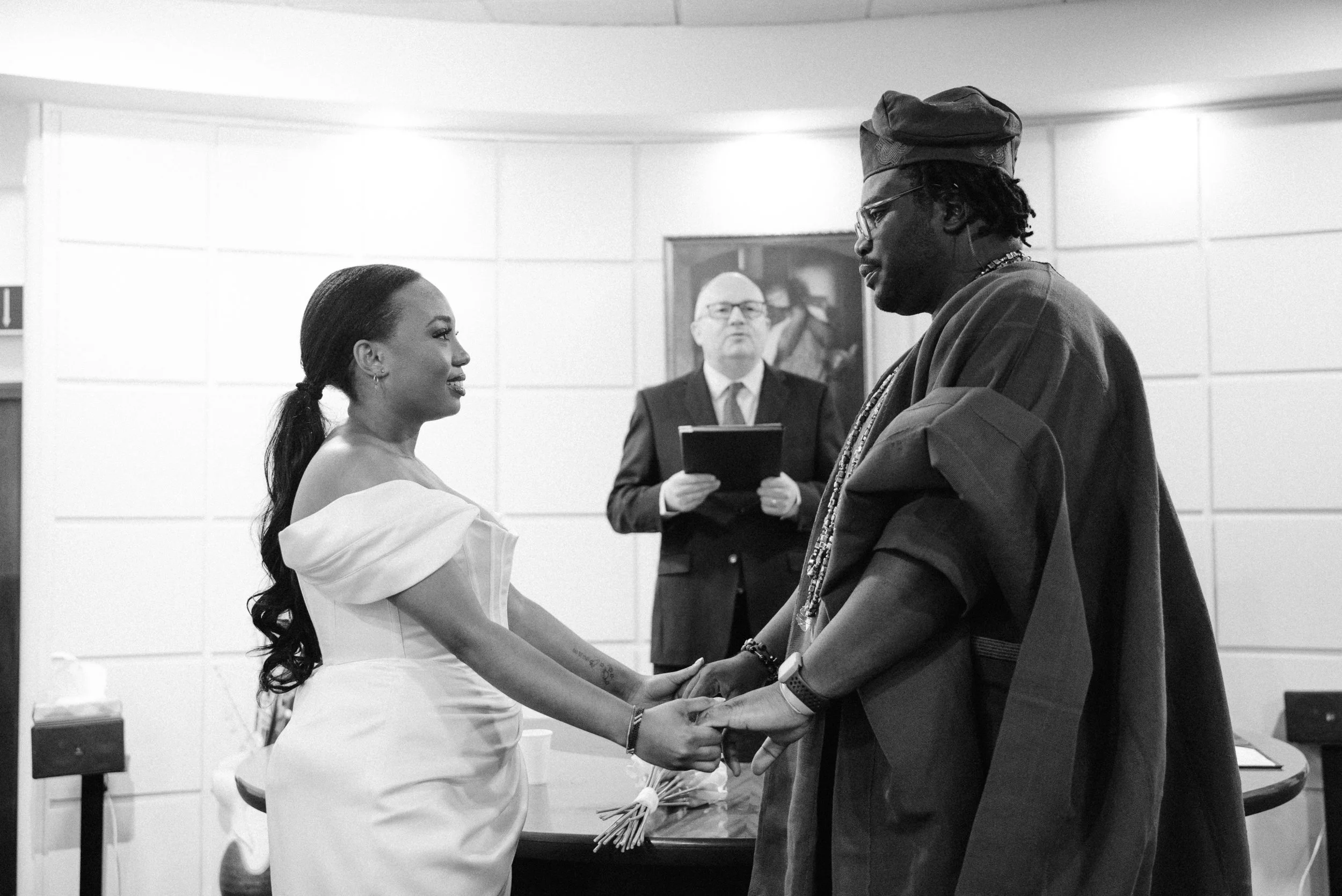 A wedding ceremony with a couple holding hands, facing each other, with an officiant standing behind them holding a notebook in a room with white paneled walls and artwork.