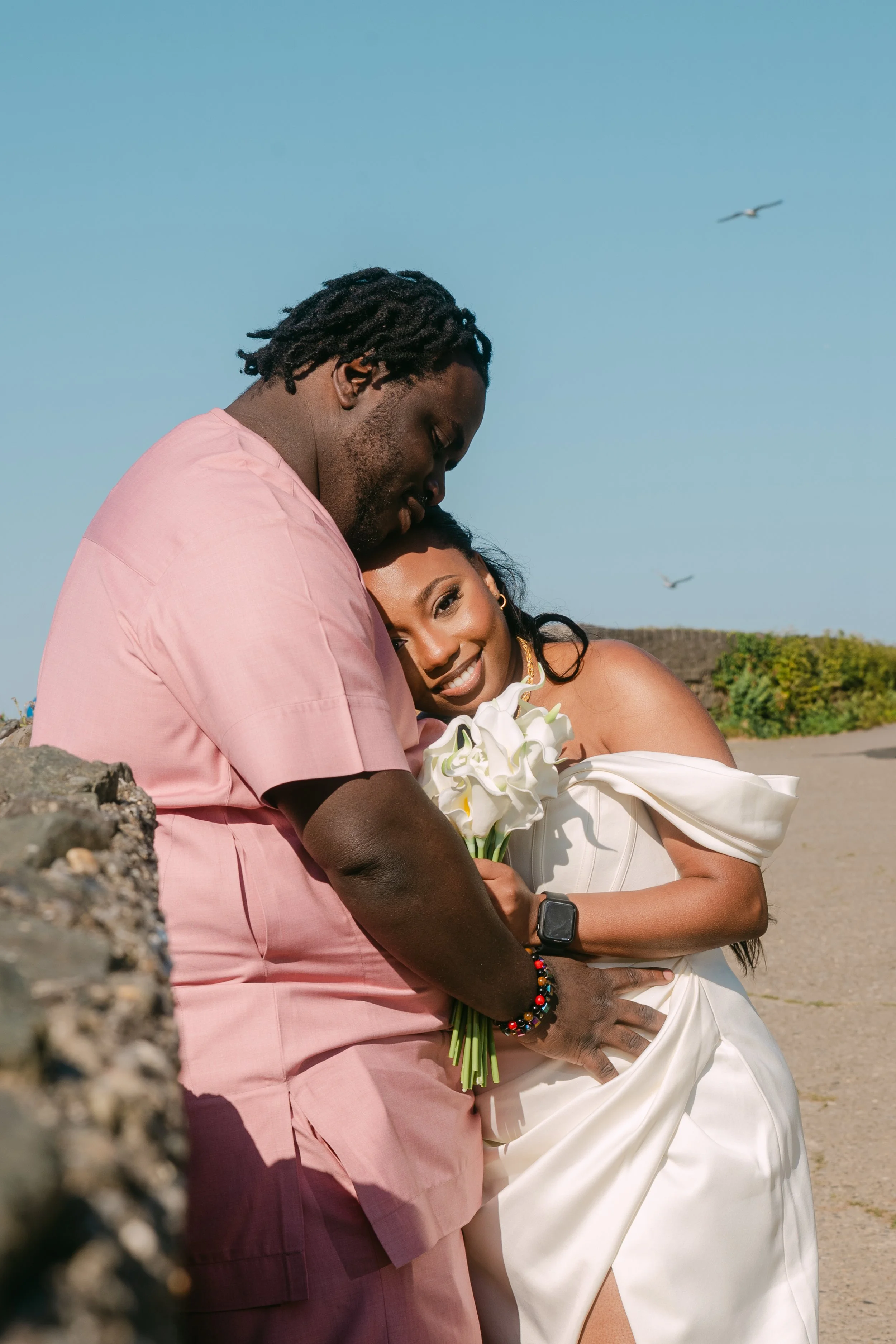 A couple on the beach with the man in pink and the woman in a white dress holding a bouquet of white flowers, embracing and smiling.