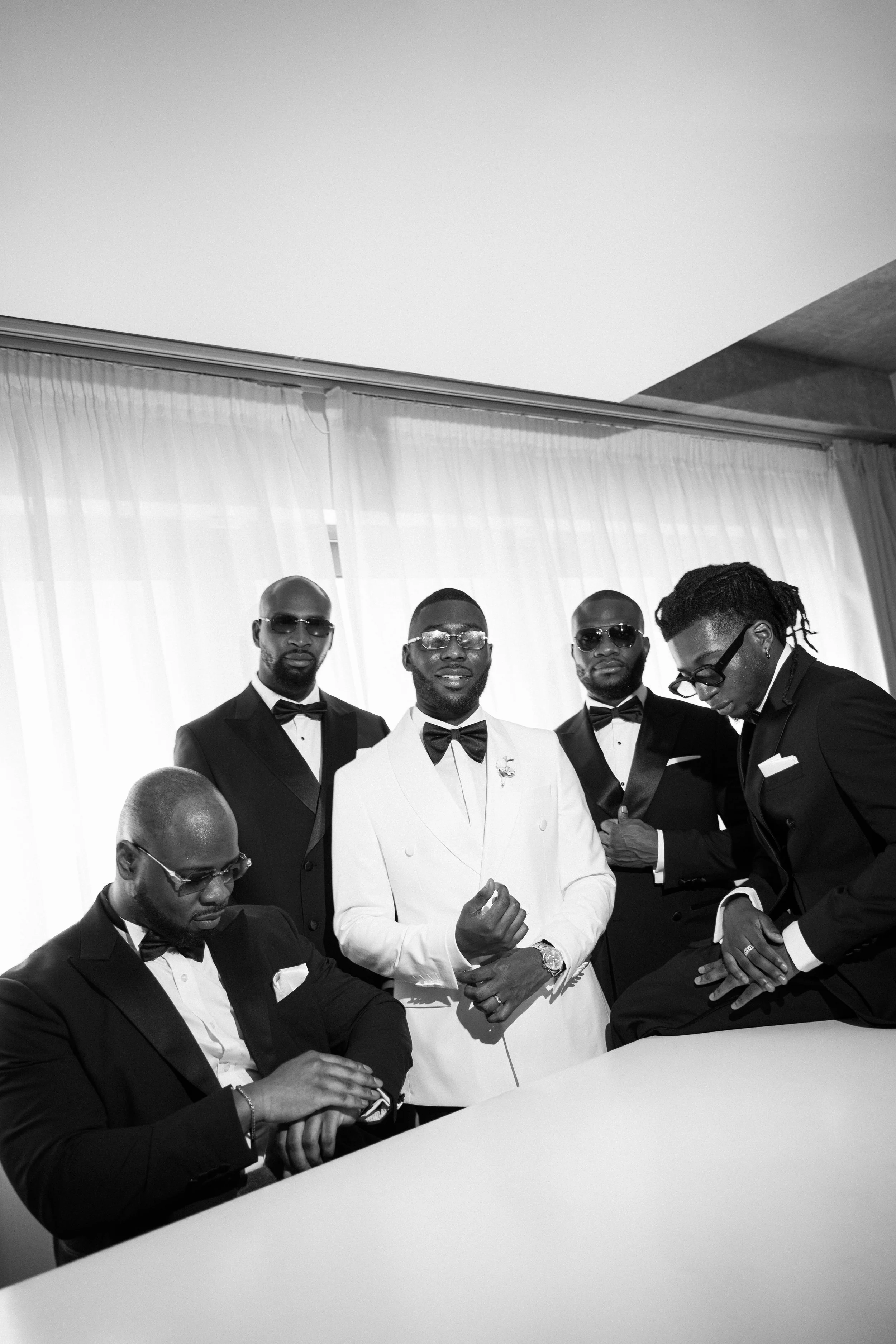 Six men dressed in tuxedos, with one in a white tuxedo jacket, posing indoors with curtains in the background.