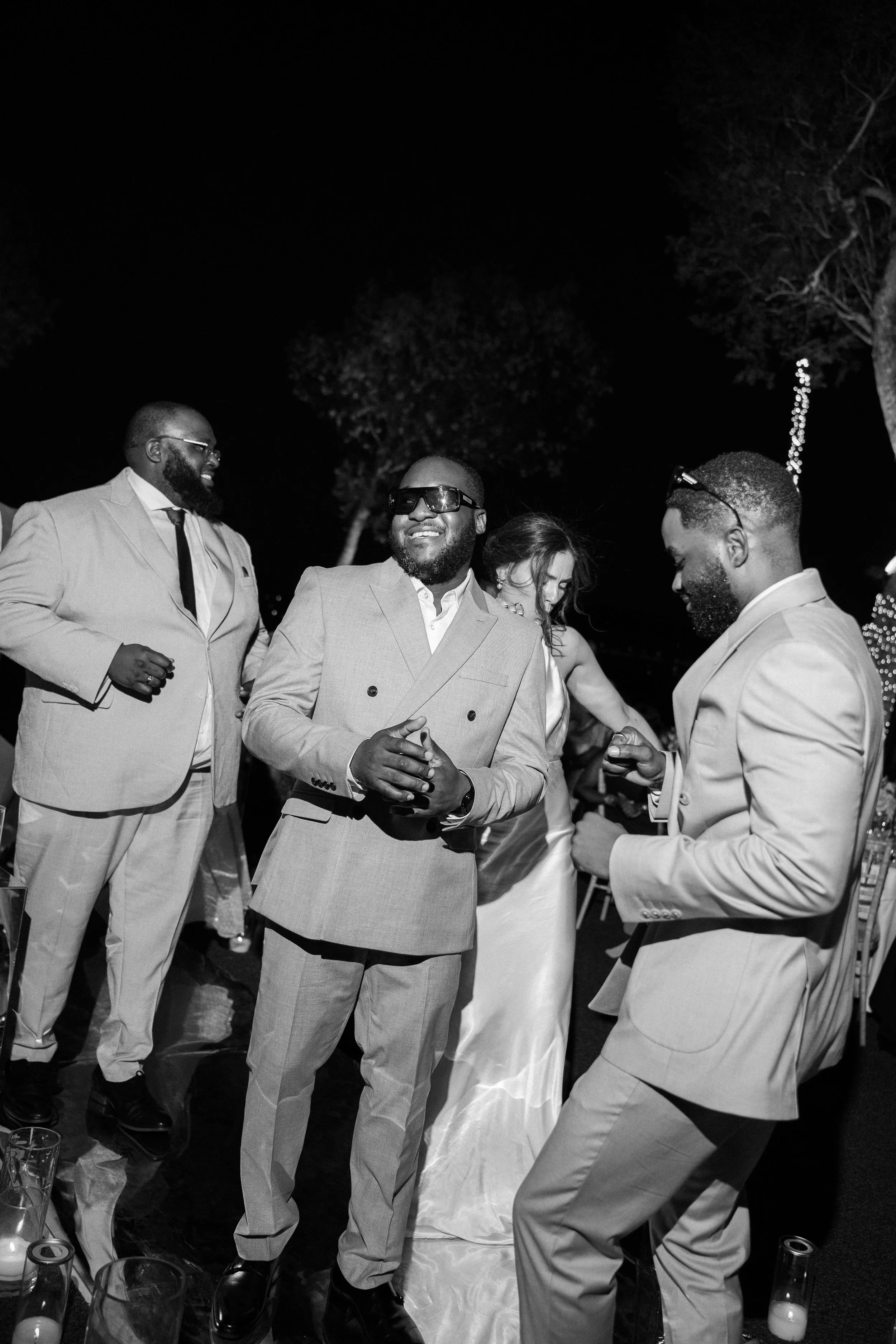 Black and white photo of five people at a celebration, dressed in suits and a dress, dancing and smiling at night outdoors with trees and decorative lights in the background.