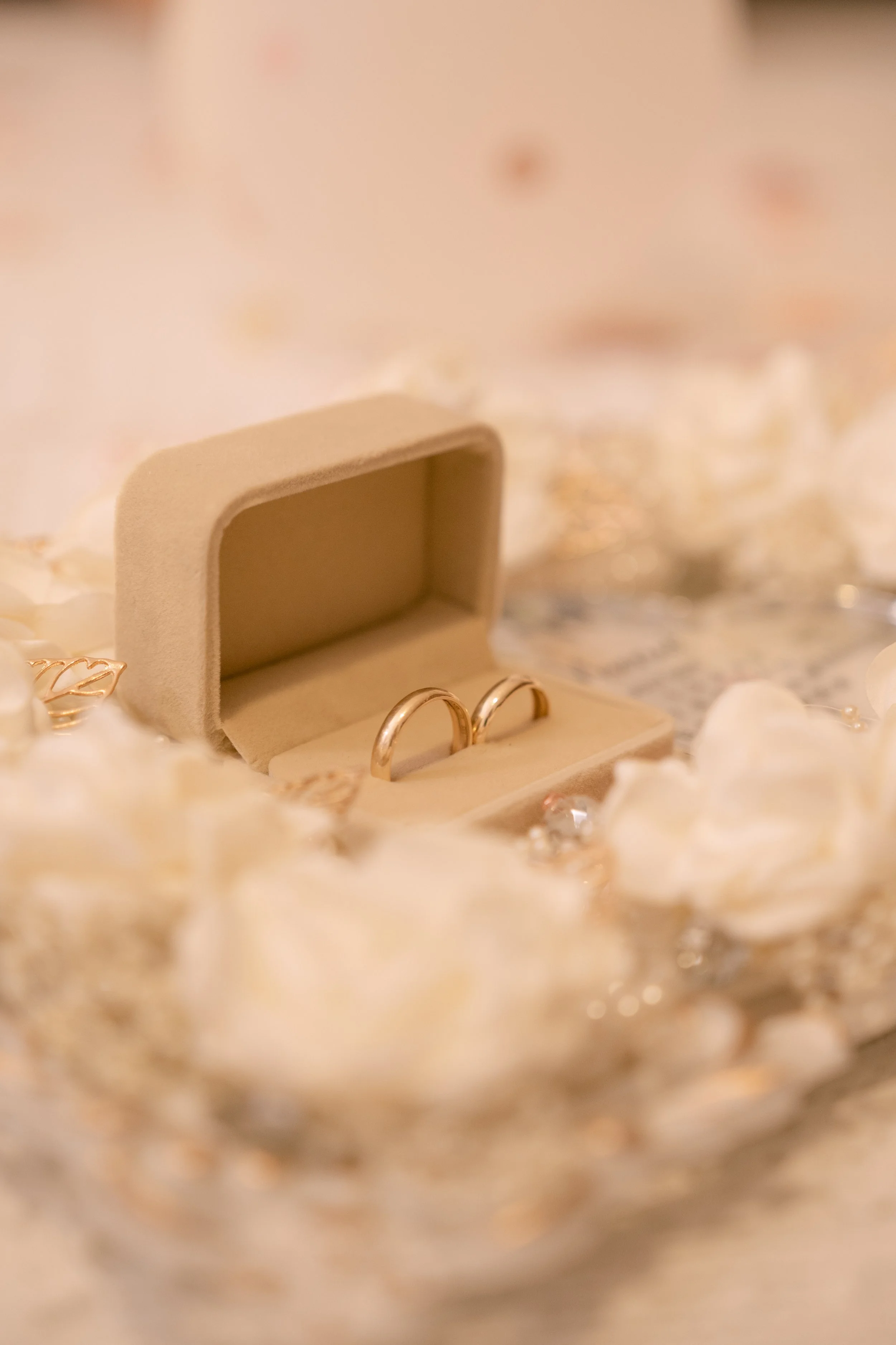 Close-up of a jewelry box with wedding rings inside, surrounded by white flower petals and sparkling decorative elements.