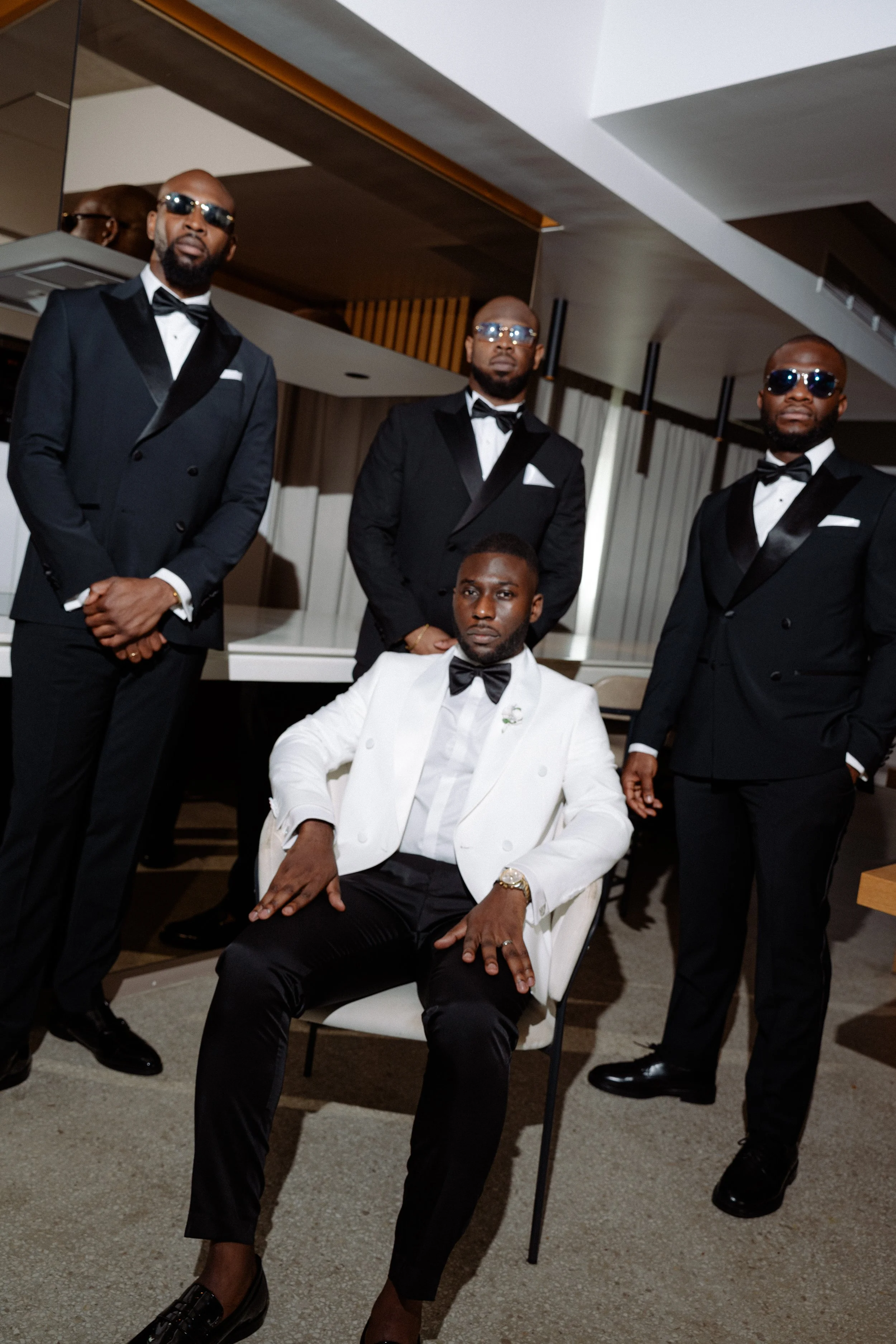 A man in a white tuxedo with a black bow tie sitting on a white chair with four men in black tuxedos standing behind him in a modern room with wooden and white accents.