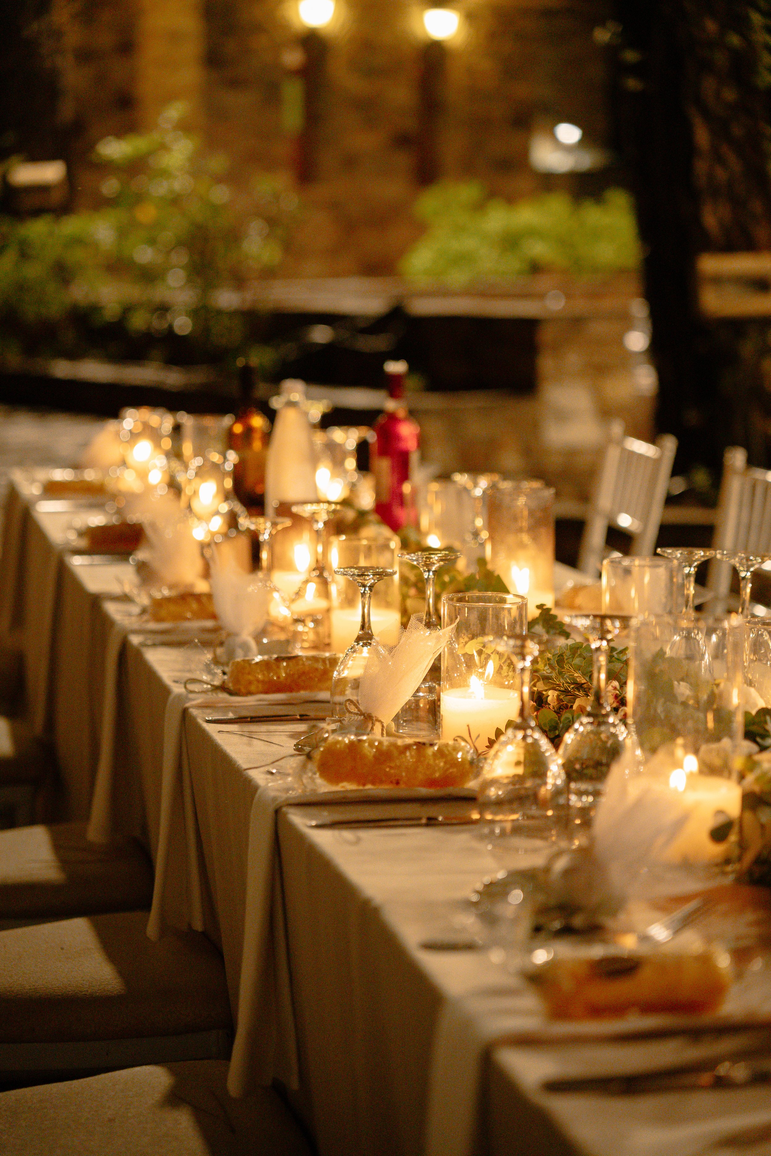 A beautifully decorated long dinner table set with white tablecloths, lit candles, glassware, and plates in a warmly lit outdoor setting at night.