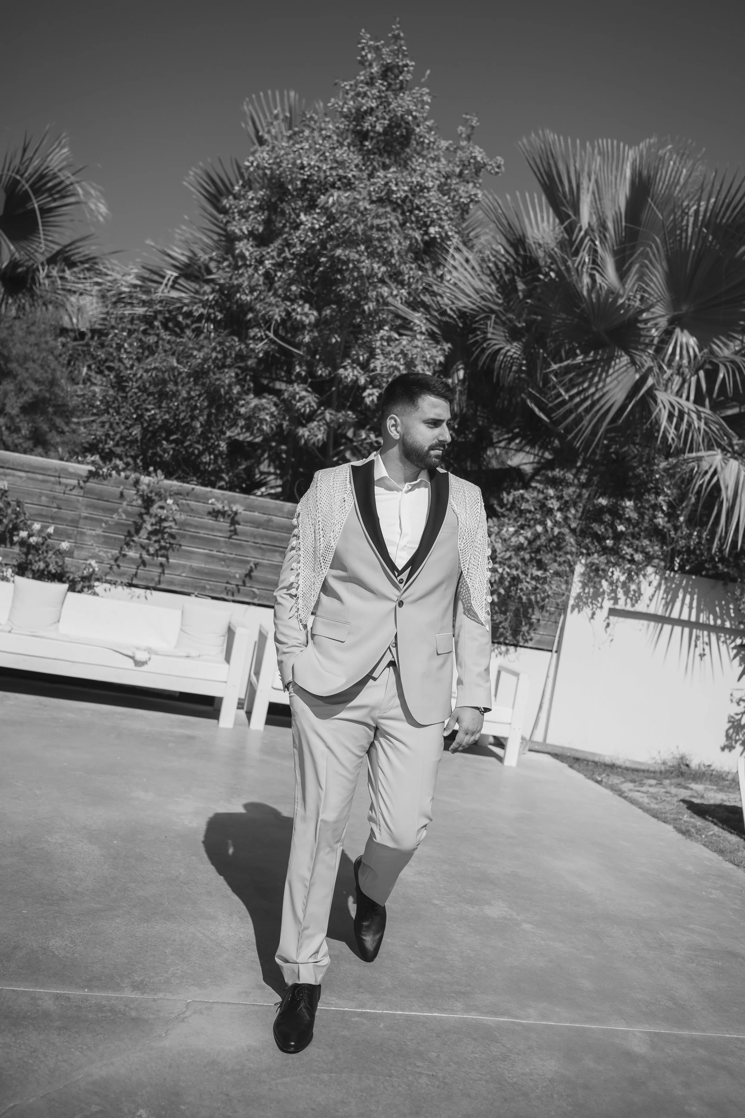 A man in a light-colored suit with a patterned blazer, walking outdoors on a sunny day, with palm trees and a bench in the background.