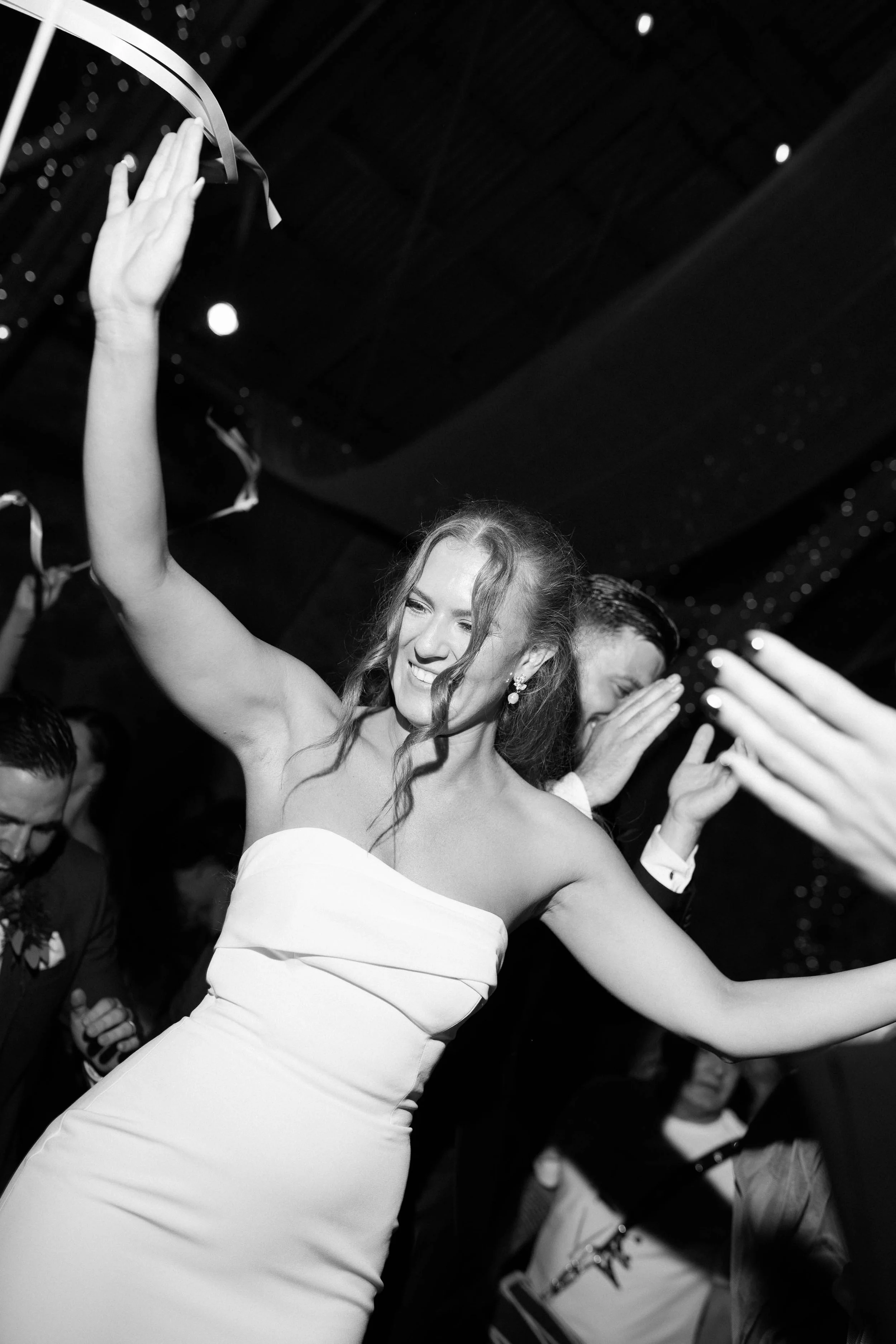 A woman in a strapless wedding dress smiling and dancing at a celebration, with others around her.