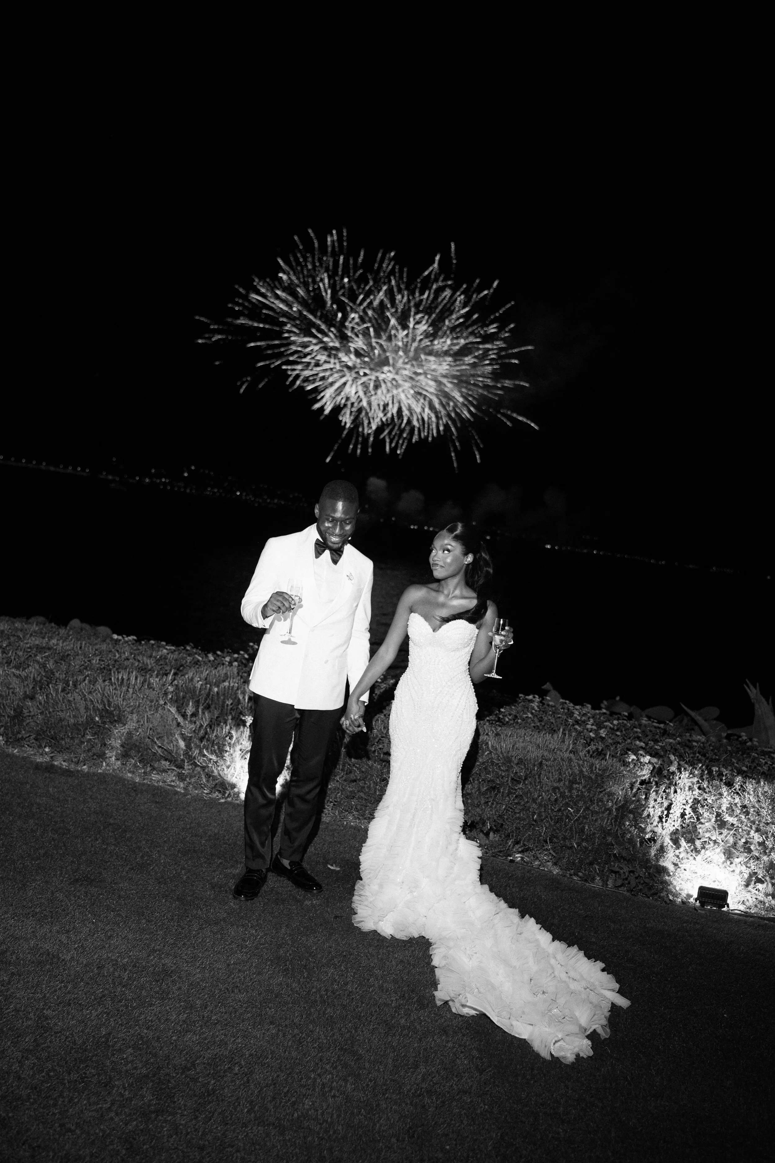 A bride and groom in wedding attire holding hands at night, with fireworks in the sky behind them, photographed in black and white.