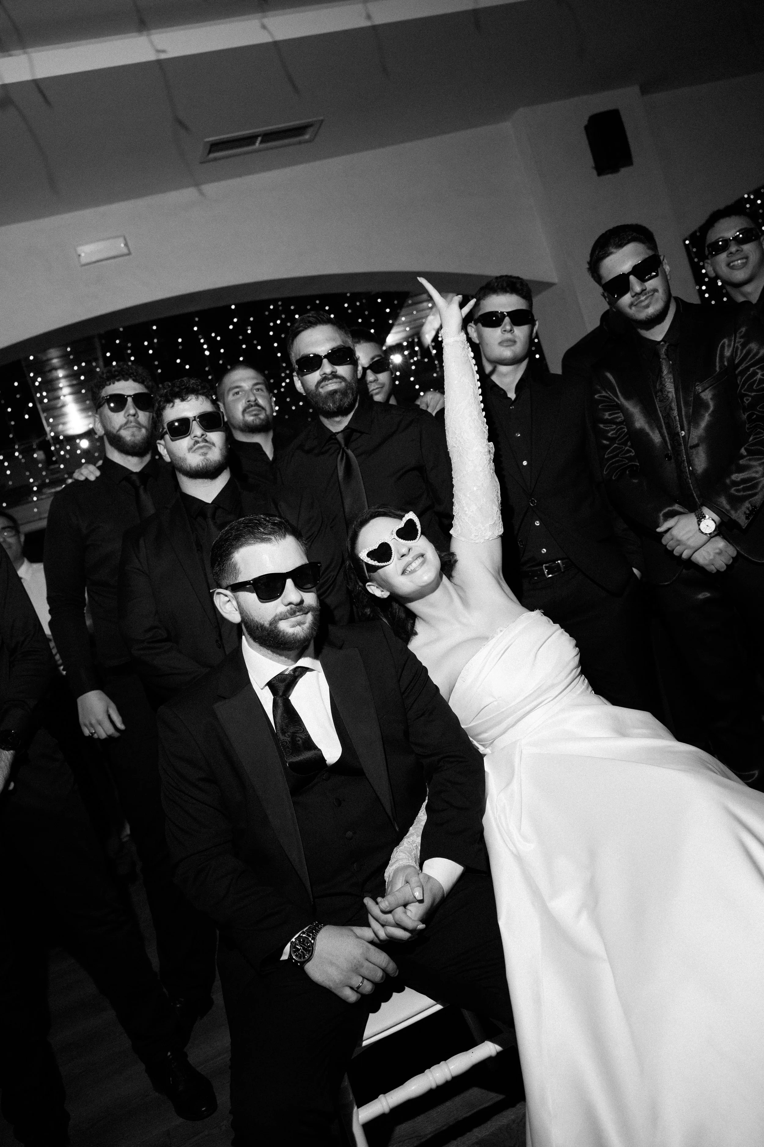 Group of people in dark clothing and sunglasses at a party, with a woman in a white dress and heart-shaped sunglasses sitting in front, smiling and raising her hand.