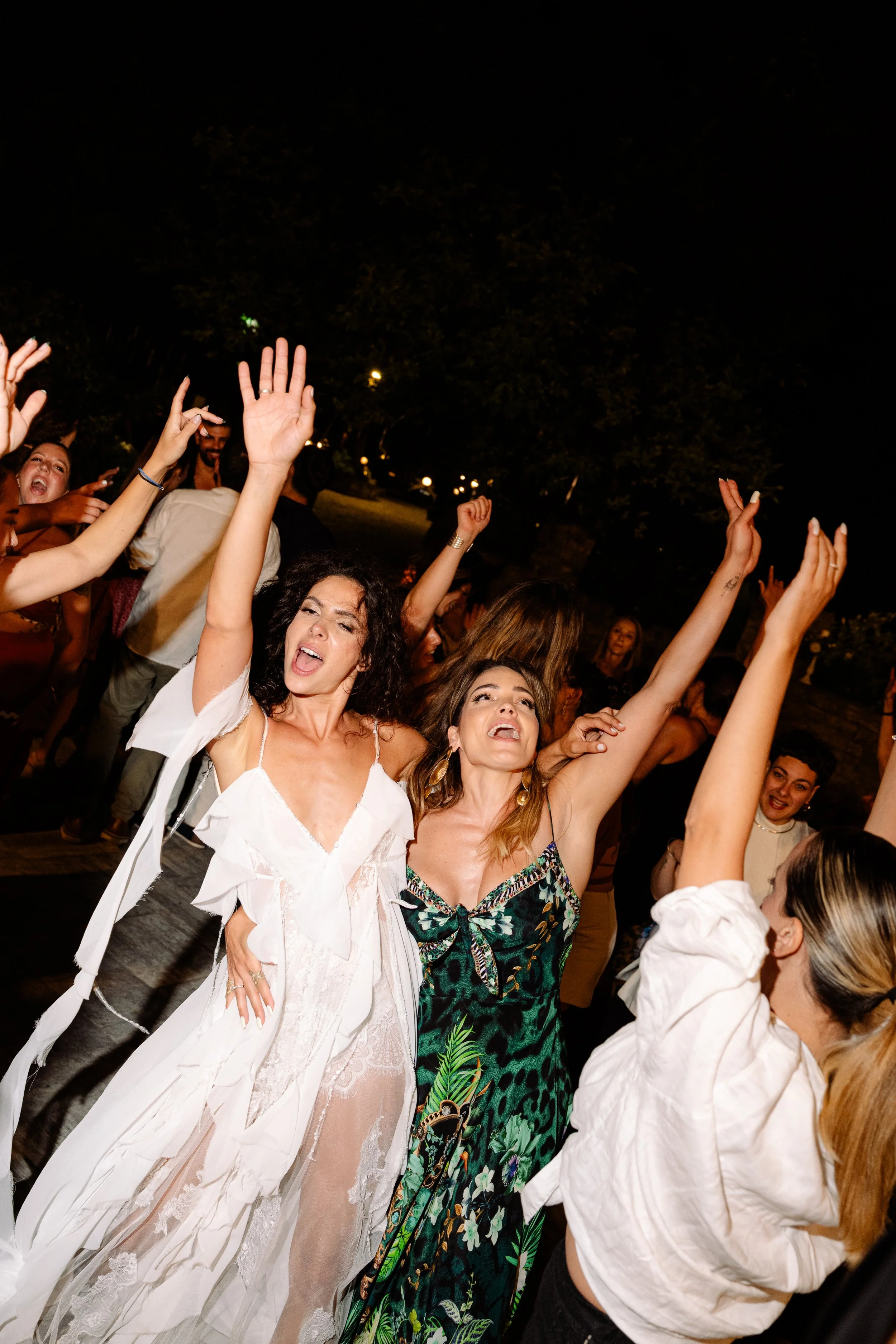 Group of people dancing and celebrating outdoors at night, some with arms raised, in a lively party scene.