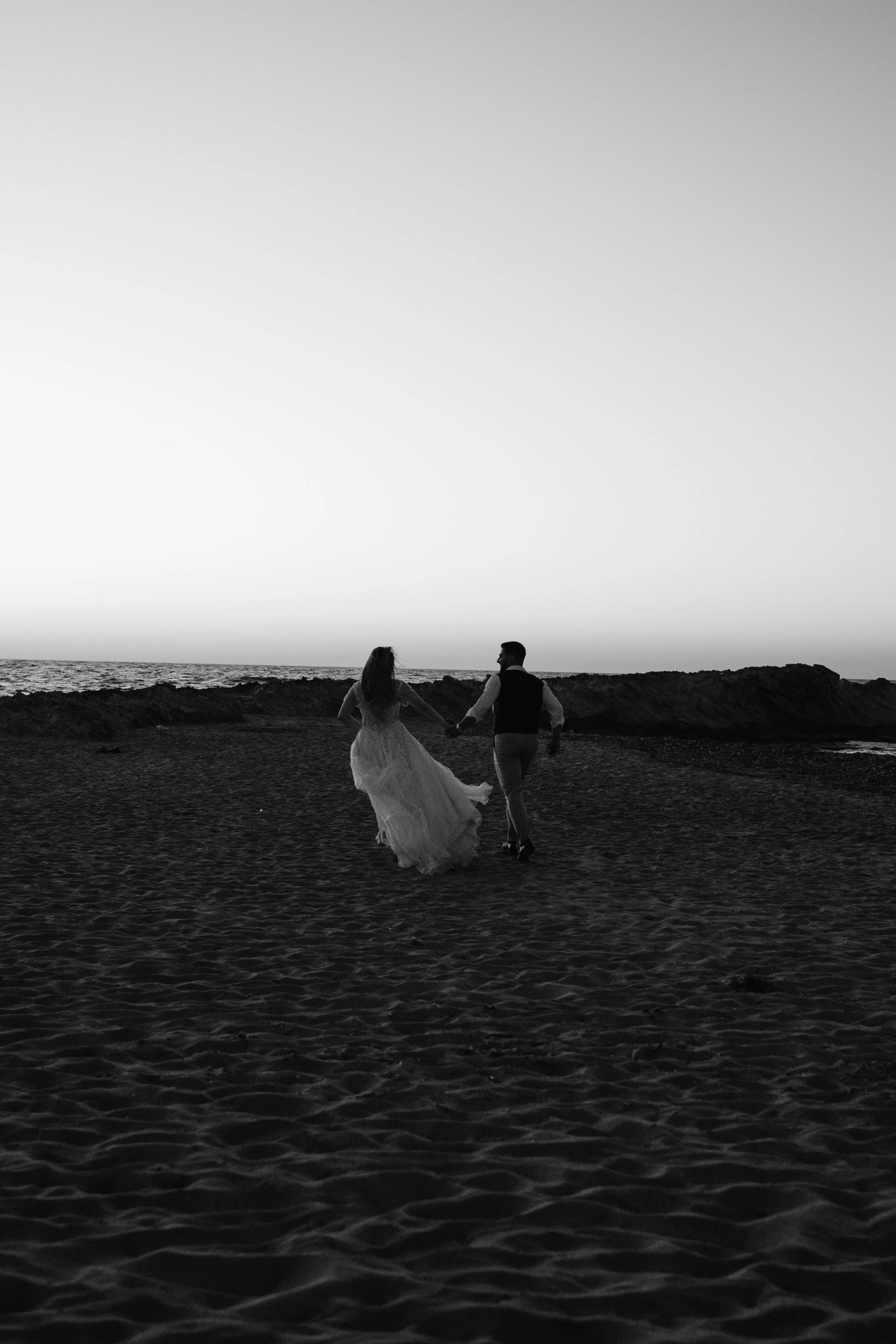 A black and white photo of a couple walking hand in hand on a beach at sunset, with the ocean in the background.