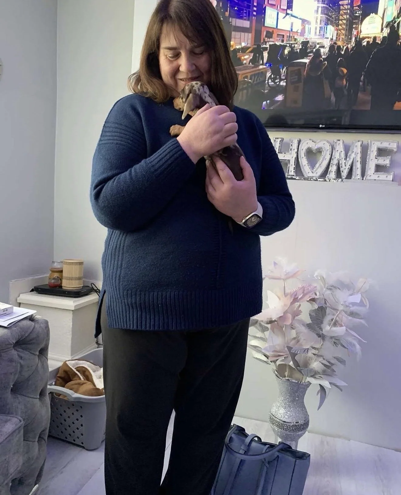 A woman holding a small puppy indoors, with a cityscape on a television screen behind her, and a decorative vase with flowers on the side.