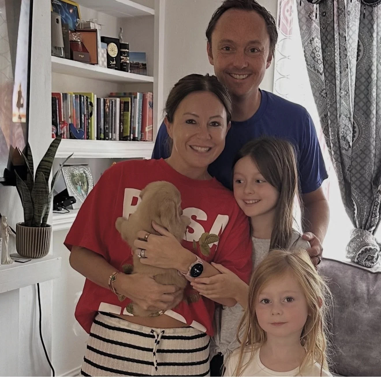 A family of five, including two adults and three children, pose indoors with a small puppy. The woman in red is holding the puppy, surrounded by the children. The man in blue stands behind them, all smiling.