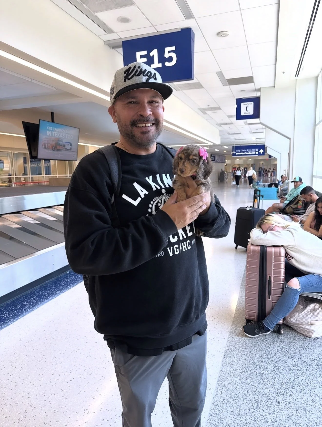 A man holding a small puppy at an airport terminal, with luggage and waiting passengers visible in the background.