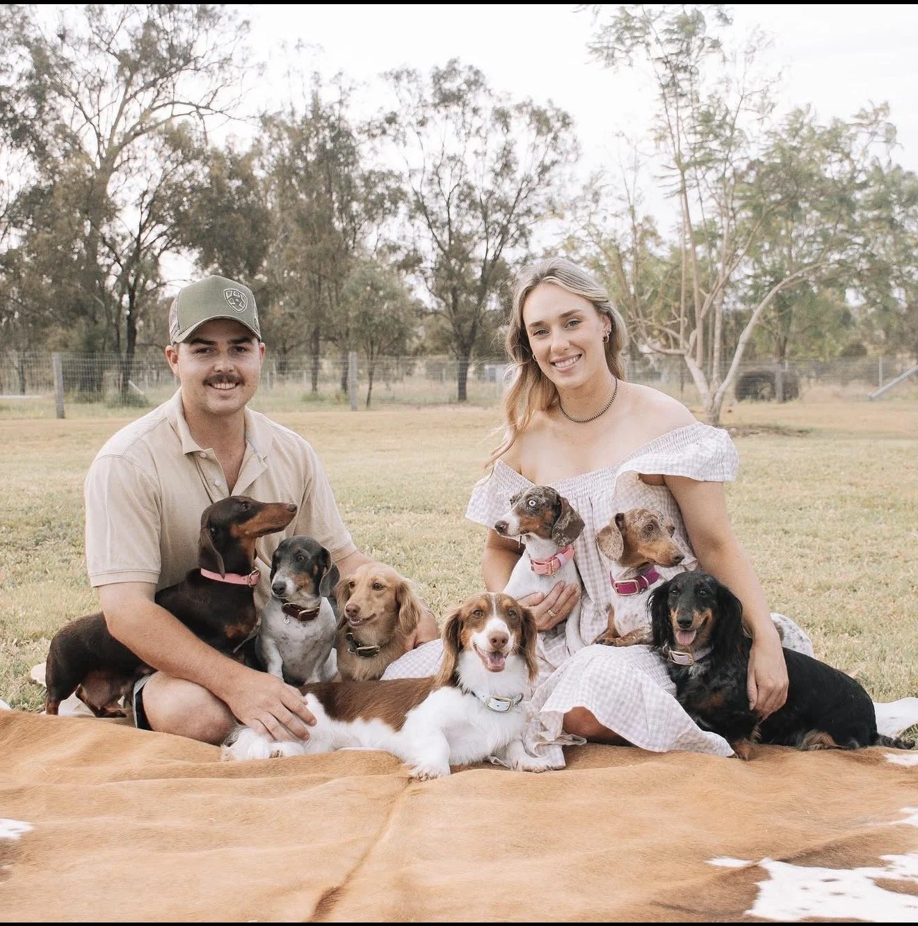 A young man and woman sitting on a blanket outdoors with eight dogs, smiling at the camera with trees and a fence in the background.