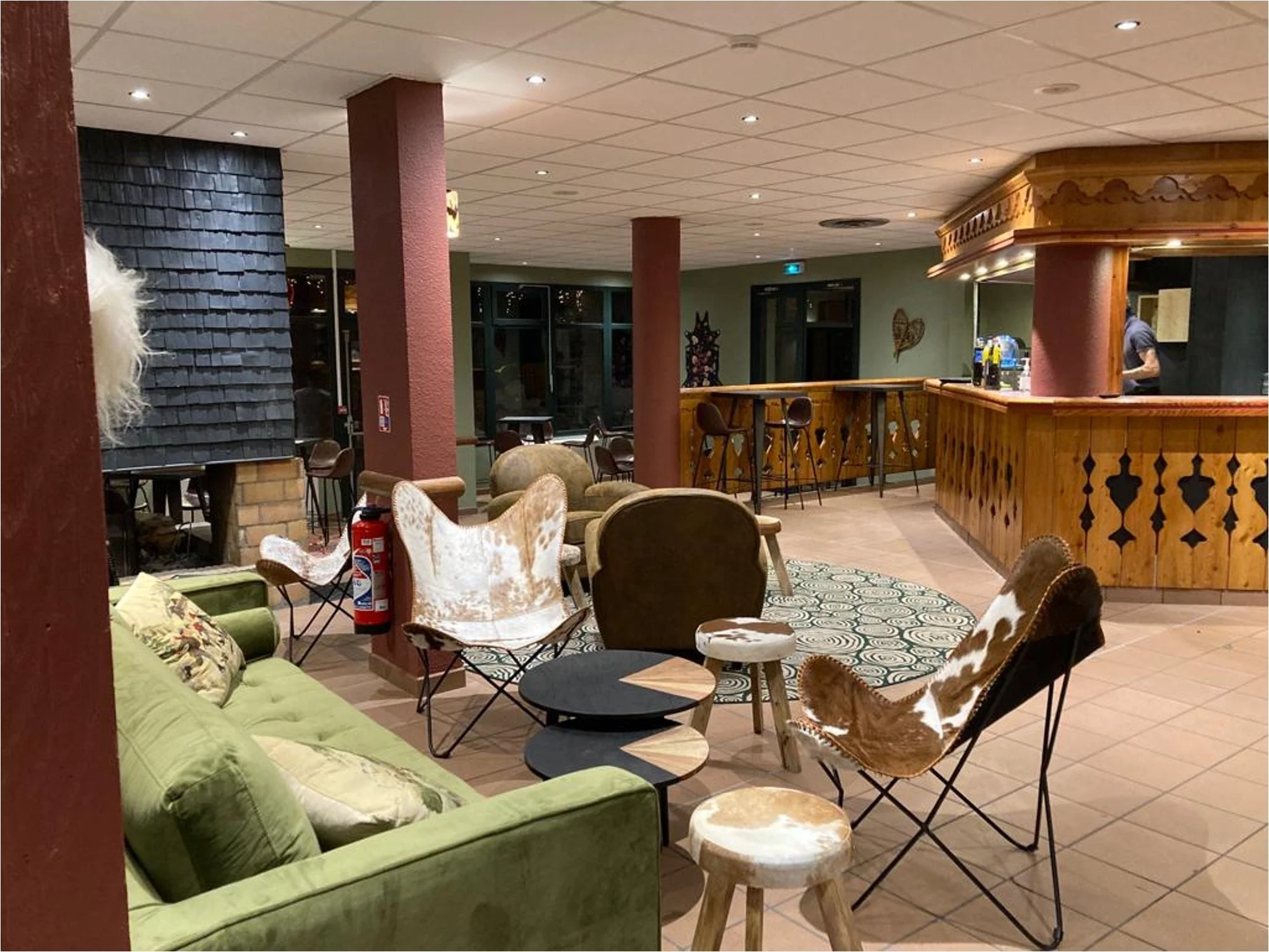 bar et salon avec un comptoir en bois, des piliers rouge, des fauteuils en peaux de vache, des fauteuils club en cuir vieilli, un canapé vert, une cheminé et un mur verre