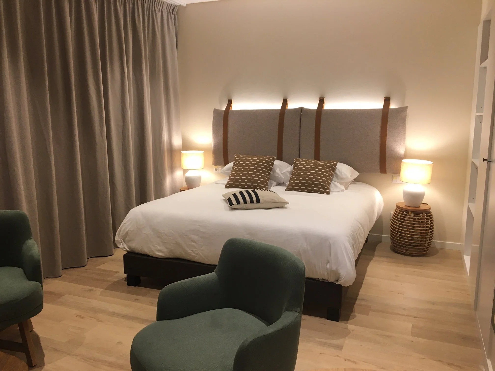 Chambre avec des rideaux gris, deux fauteuils vert, deux lampes de chevet, un lit blanc avec des coussins et une tête de lit.