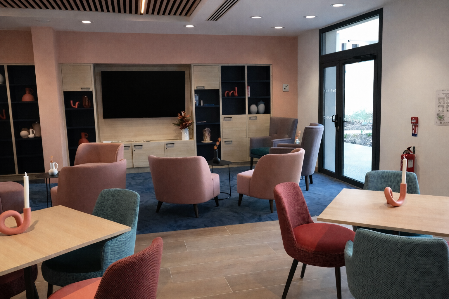 Salon moderne avec des chaises colorées rouge, bleu et rose saumon. Un tapis bleu et une télévision accrochée dans une bibliothèque en bois