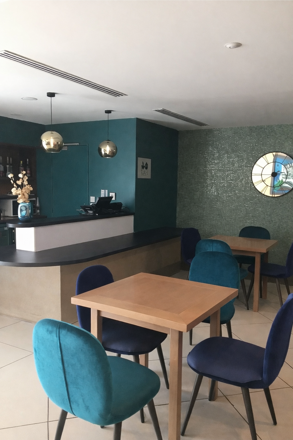 Comptoir de bar avec pot de fleur, luminaires circulaires et un mur bleu. Des tables en bois et des chaises bleu. Une horloge sur un mur vert