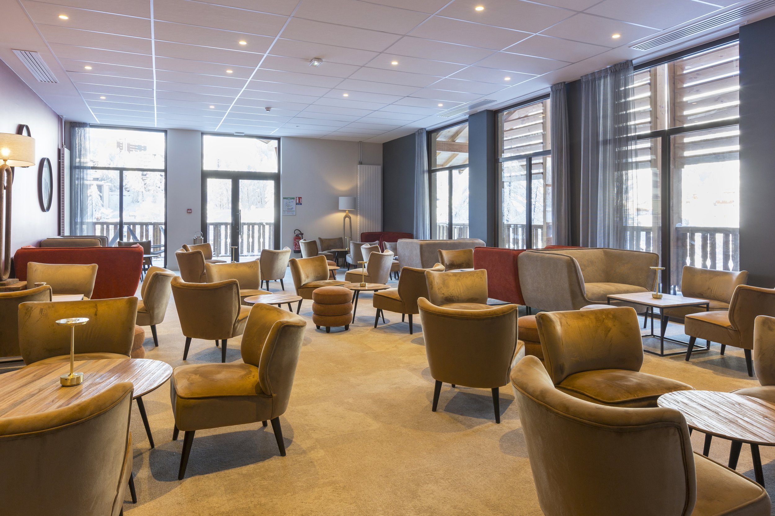 Salon d'hôtel avec des chaises basses beige, des tables basses en bois, des miroirs accrochés sur un mur blanc et plusieurs baies vitrées.